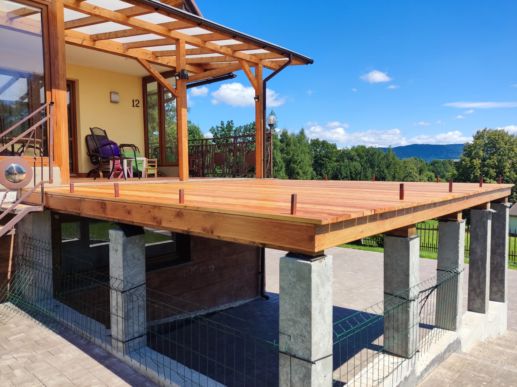 Nowoczesny taras z zadaszeniem i drewnianą podłogą, wsparty na betonowych słupach, z widokiem na zielony krajobraz w słoneczny dzień.