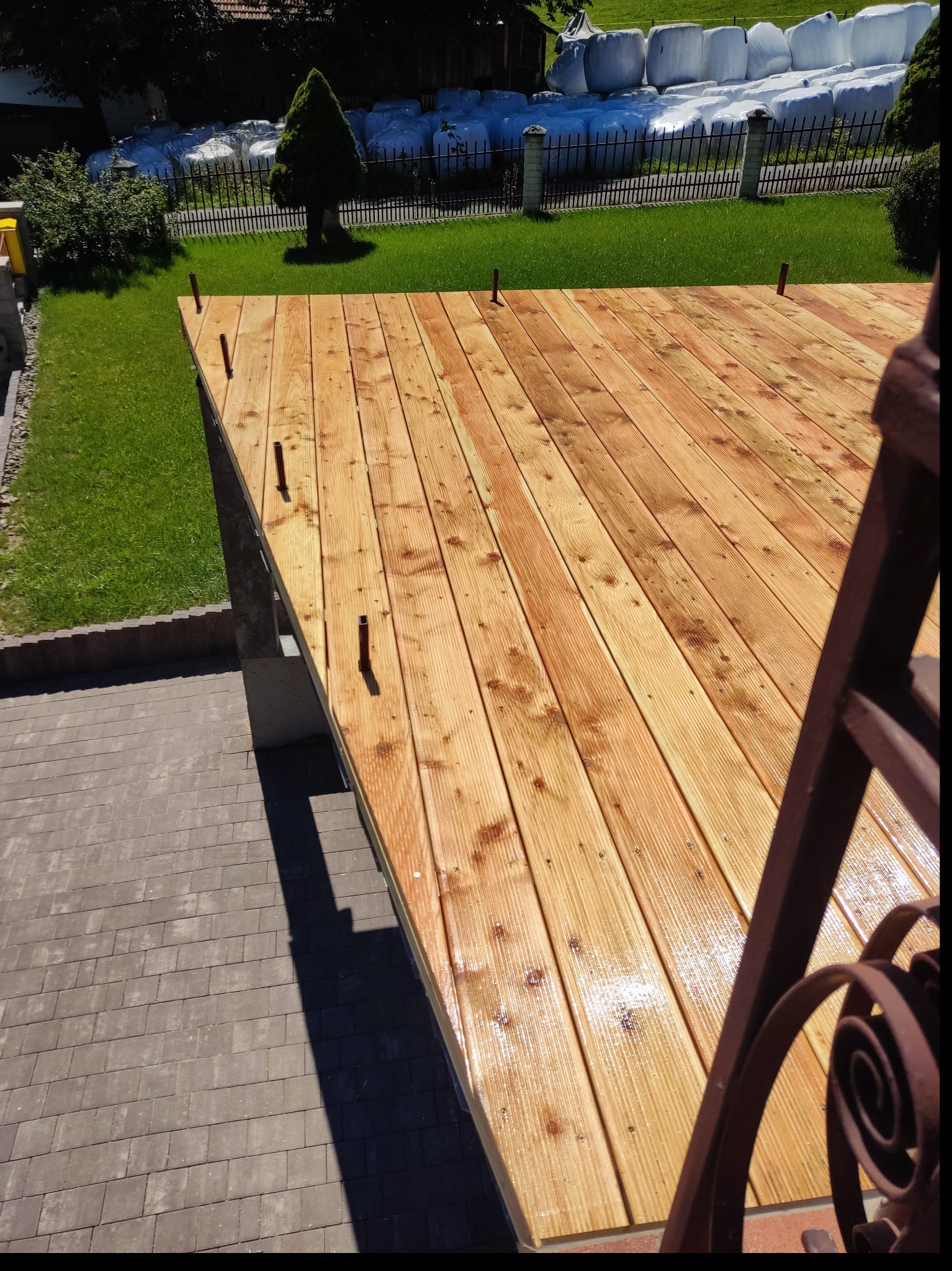 Świeżo zaimpregnowany, jasny, drewniany podest tarasu z wystającymi elementami montażowymi, widok z góry na tle trawnika i białych bel słomy.