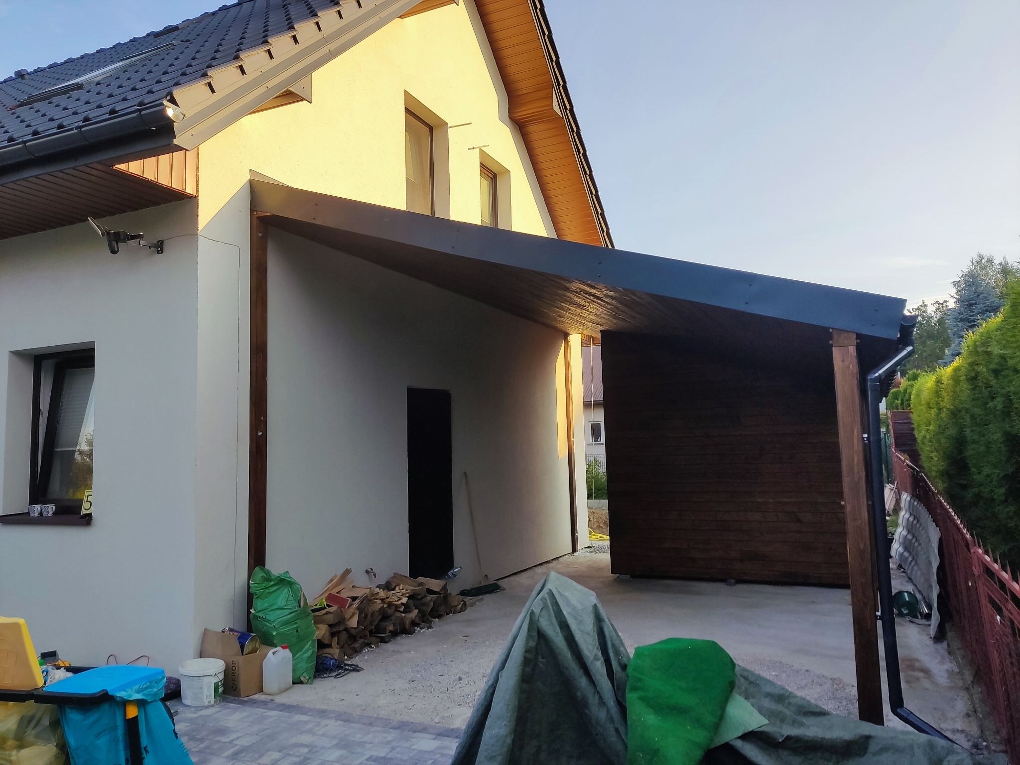 Jednospadowa wiata samochodowa z szopą narzędziową, przymocowana do ściany domu, widok z zewnątrz na tle budynku mieszkalnego i zieleni.