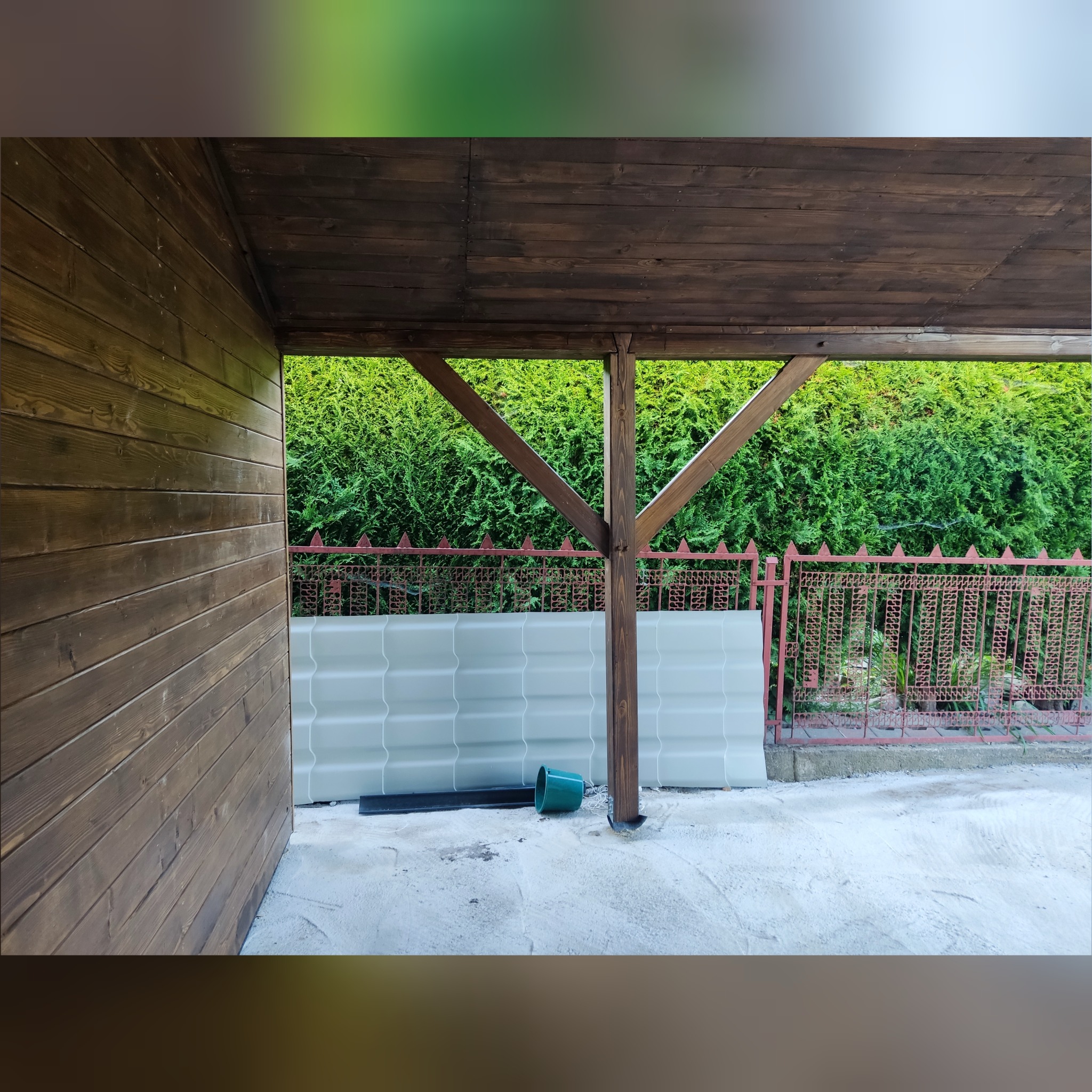 Wnętrze jednospadowej wiaty samochodowej z drewnianą konstrukcją, widoczne przygotowane płyty do zabudowy oraz szopa narzędziowa w tle.