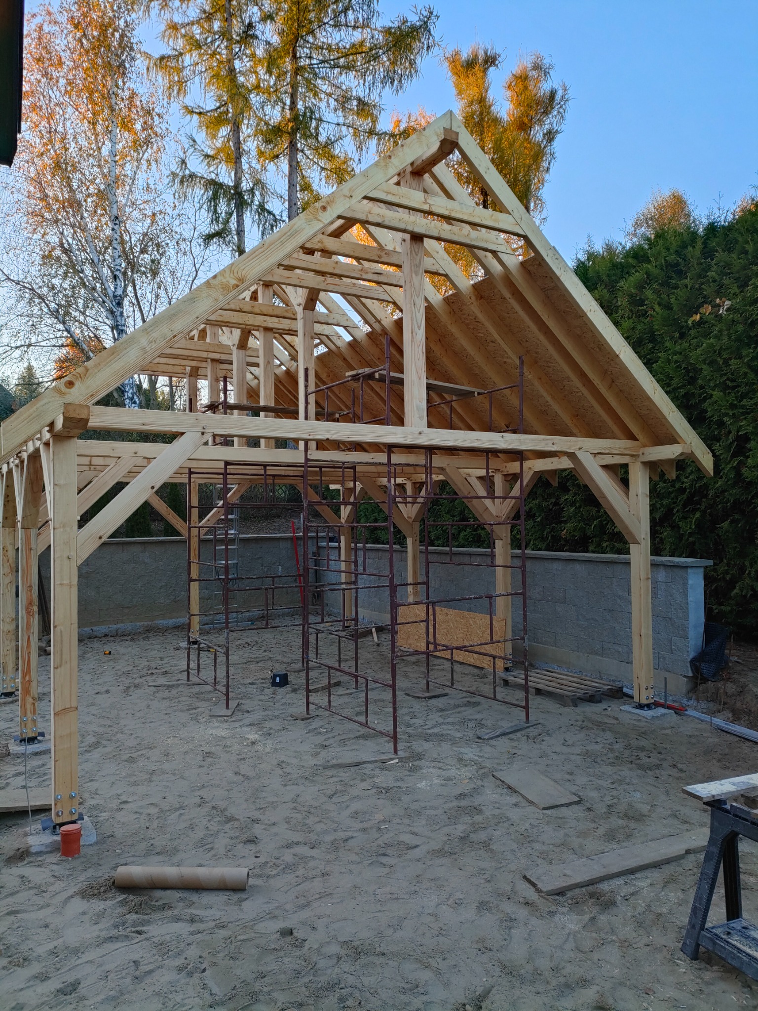 Konstrukcja z jasnego drewna w trakcie budowy, widoczne rusztowania i elementy mocujące. W tle drzewa i fragment muru.