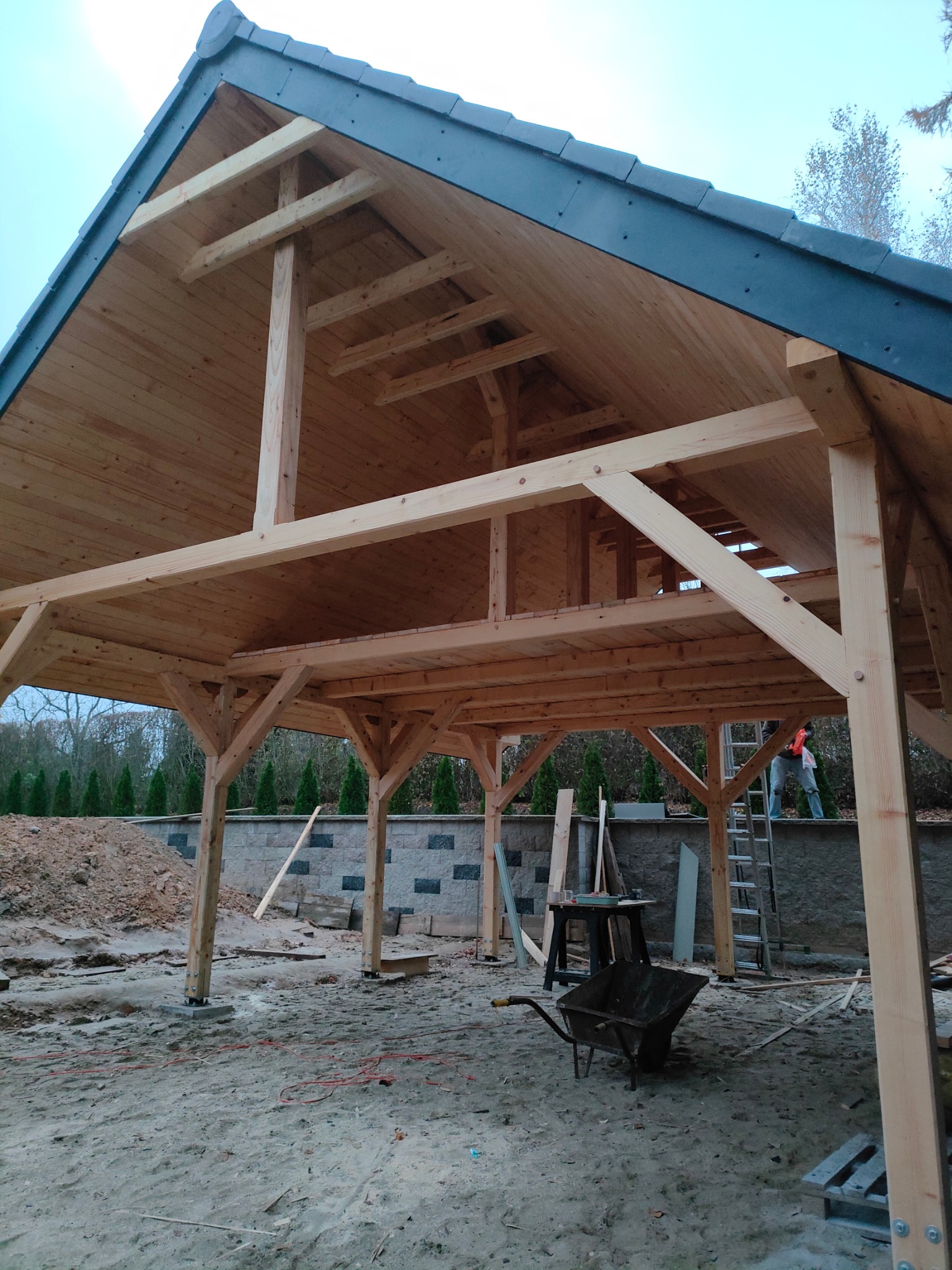 Wnętrze konstrukcji z jasnego drewna z ciemnym dachem, w trakcie budowy. Widoczne elementy szkieletu, słupy, podłoże z piasku, taczka i drabina. Prace w toku.