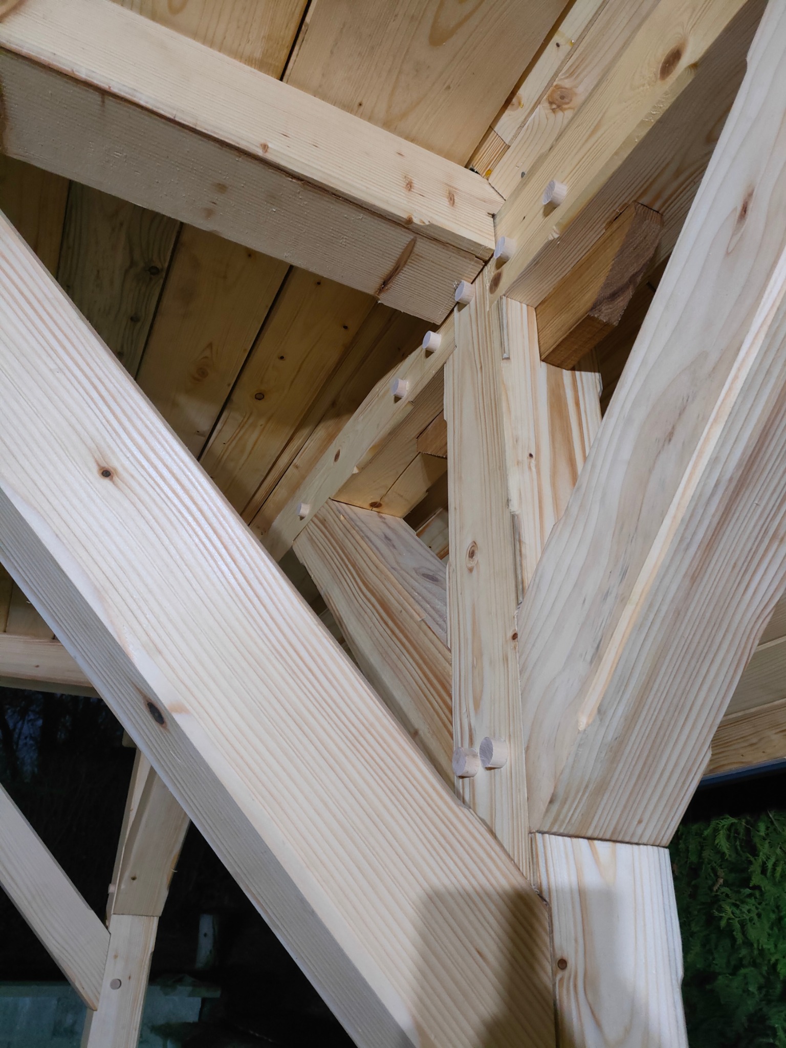 Konstrukcja z jasnego drewna, widoczne połączenia na kołki, surowe drewno, detale ciesielskie, widok z dołu na drewniany szkielet dachu lub altany w trakcie budowy.