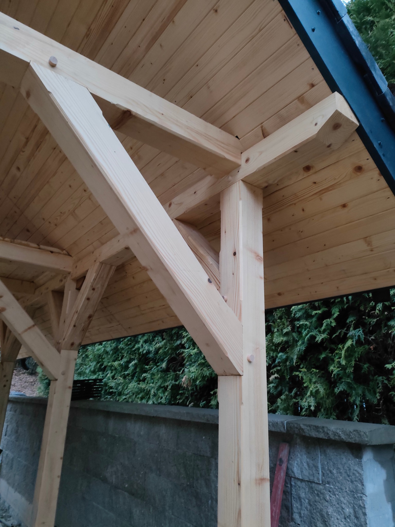 Surowe drewno tworzy konstrukcję zadaszenia, widoczne połączenia na kołki, tło z zielonych krzewów i kamienny murek. Ujęcie od dołu, podkreślające solidność wykonania.