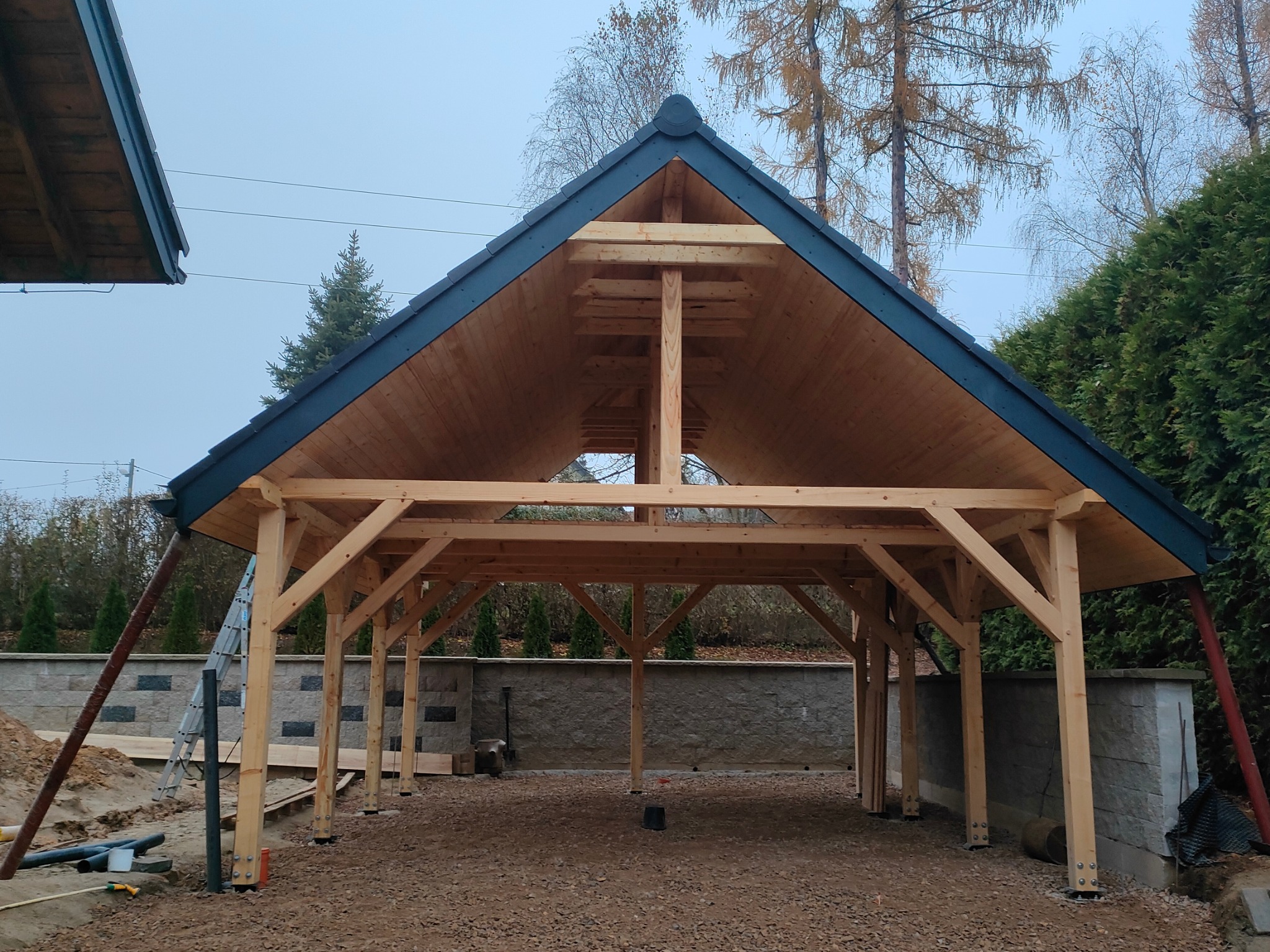 Konstrukcja z jasnego drewna z ciemnym dachem w ogrodzie, widok z przodu. Elementy montażowe widoczne u podstawy słupów. W tle drzewa i krzewy.