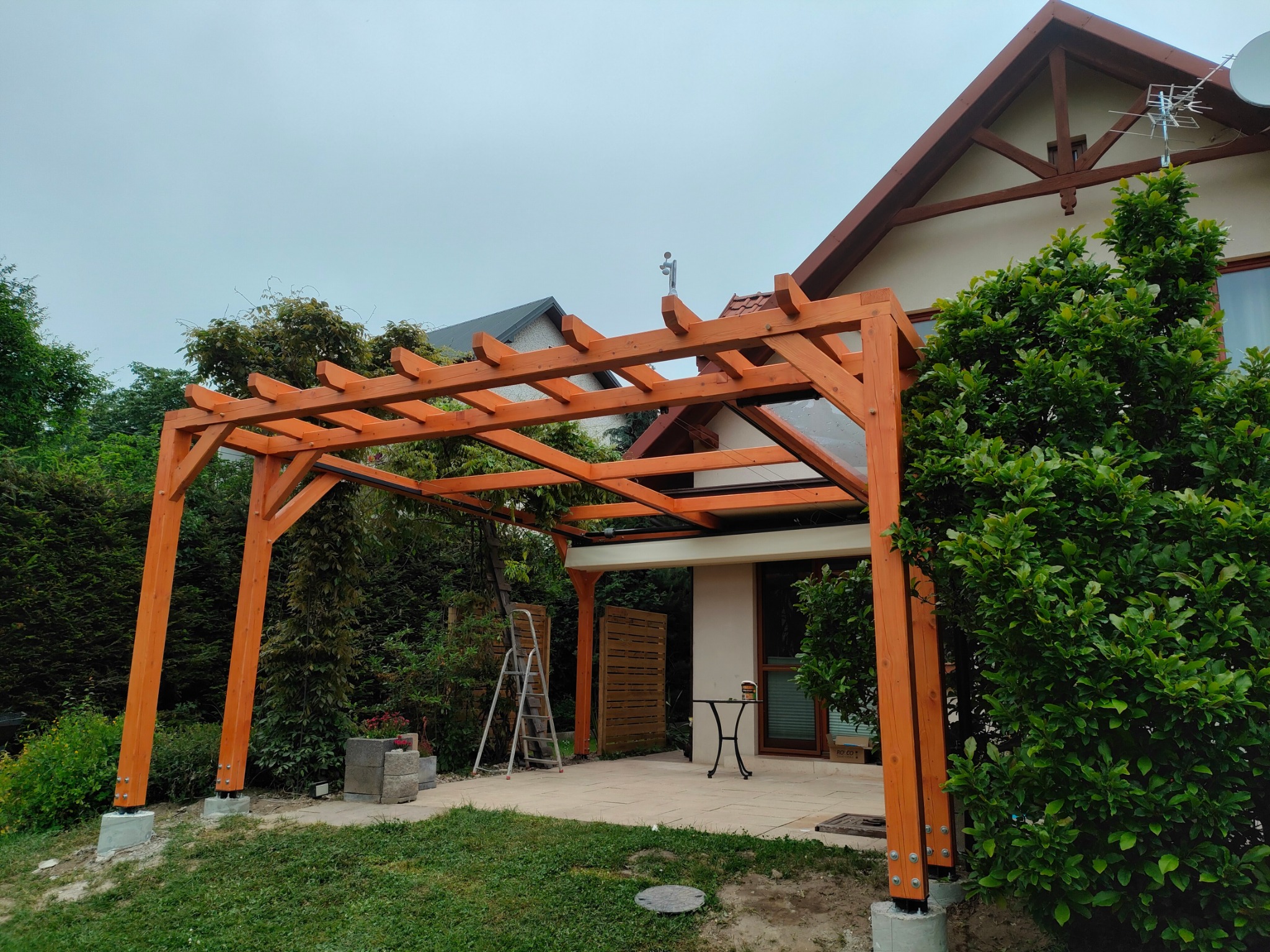 Drewniana pergola z przezroczystym zadaszeniem, widoczna z zewnątrz, z elementami mocującymi. W tle dom z ogrodem i drabina oparta o konstrukcję.