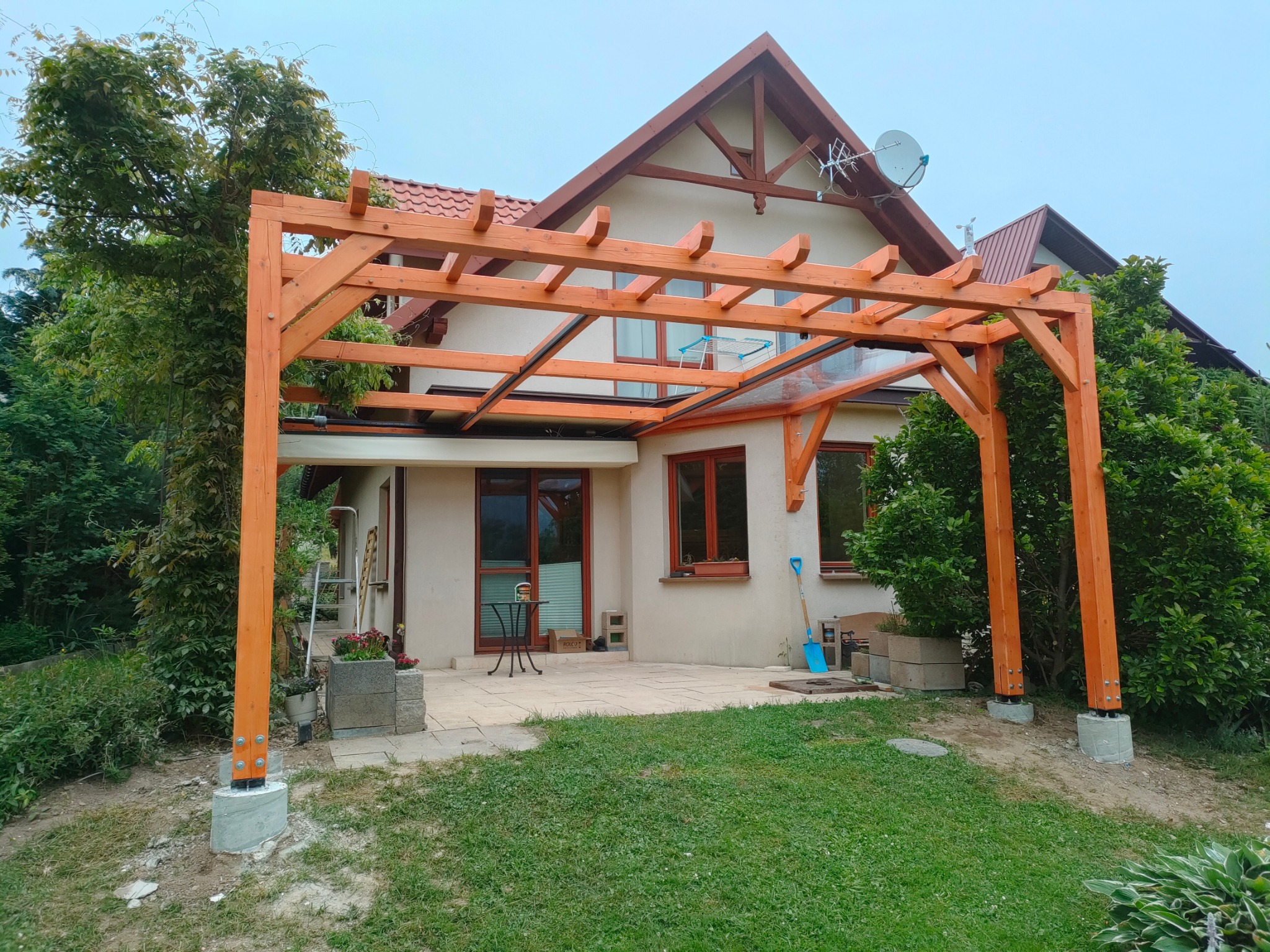 Jasna, drewniana pergola z przezroczystym zadaszeniem, częściowo zacieniona przez zieleń, przylegająca do domu z beżową elewacją i brązowymi oknami. Widok z ogrodu.