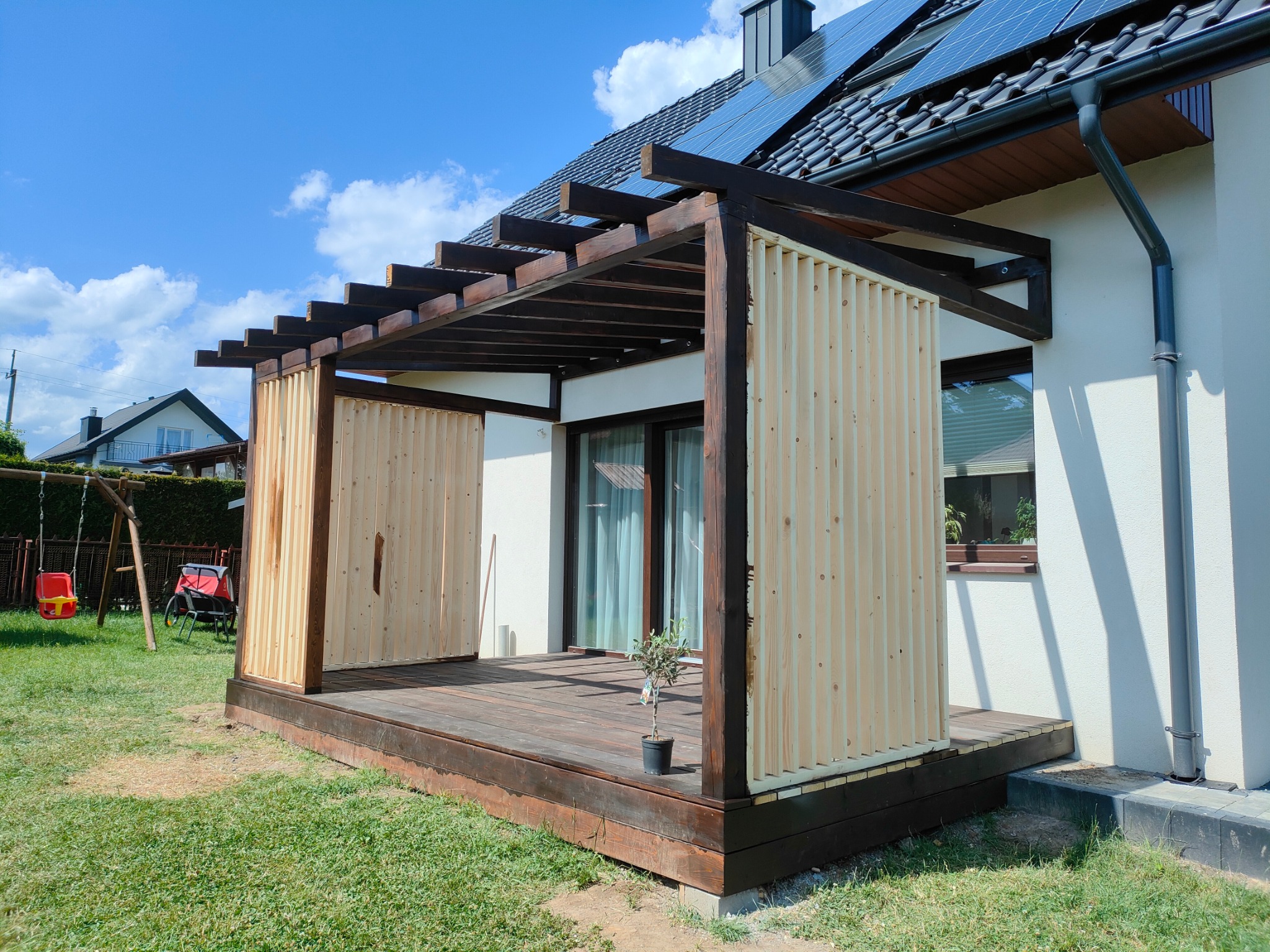 Nowoczesna konstrukcja z drewna z jasnymi lamelami i ciemnym szkieletem, tworząca zacieniony taras przy domu z panelami słonecznymi na dachu. Małe drzewko w doniczce na podłodze.