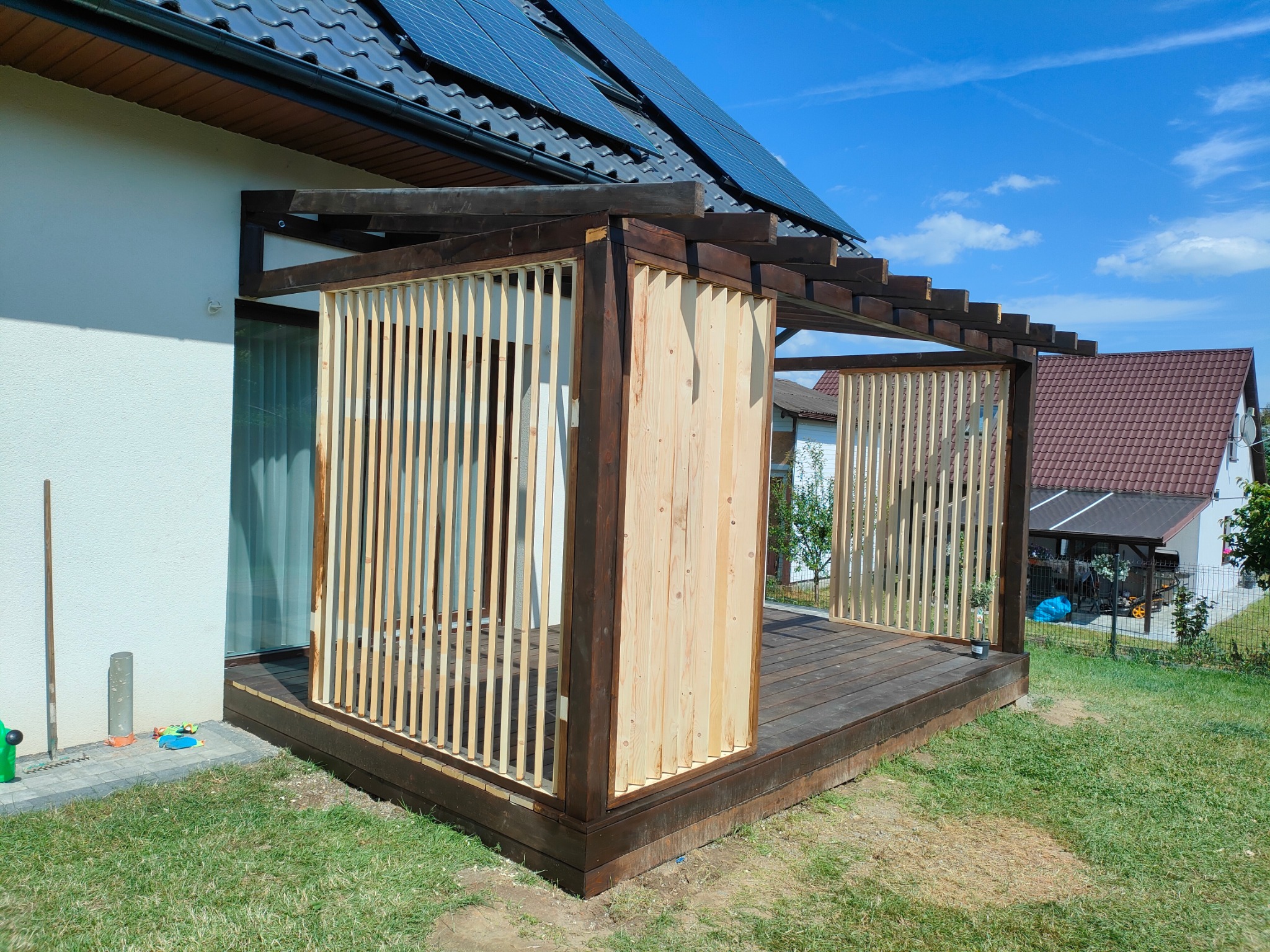 Nowoczesna konstrukcja tarasowa z drewnianymi lamelami, częściowo osłonięta, z widocznym fragmentem domu z panelami słonecznymi i zielonym trawnikiem.