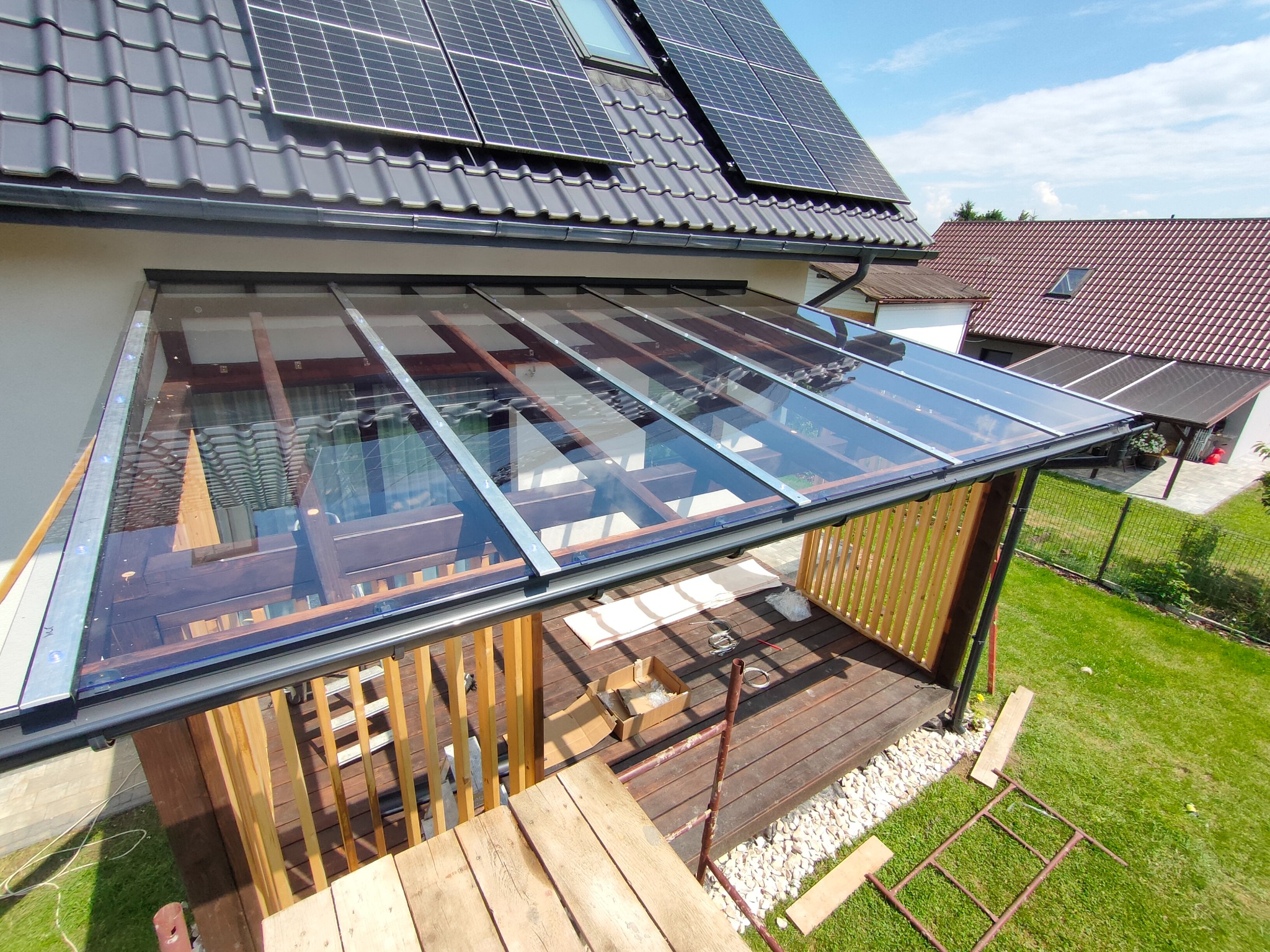 Szklana zabudowa tarasu z drewnianą podłogą w trakcie montażu, widok z góry. Na dachu domu panele słoneczne. W tle dom z czerwoną dachówką i zielony trawnik.