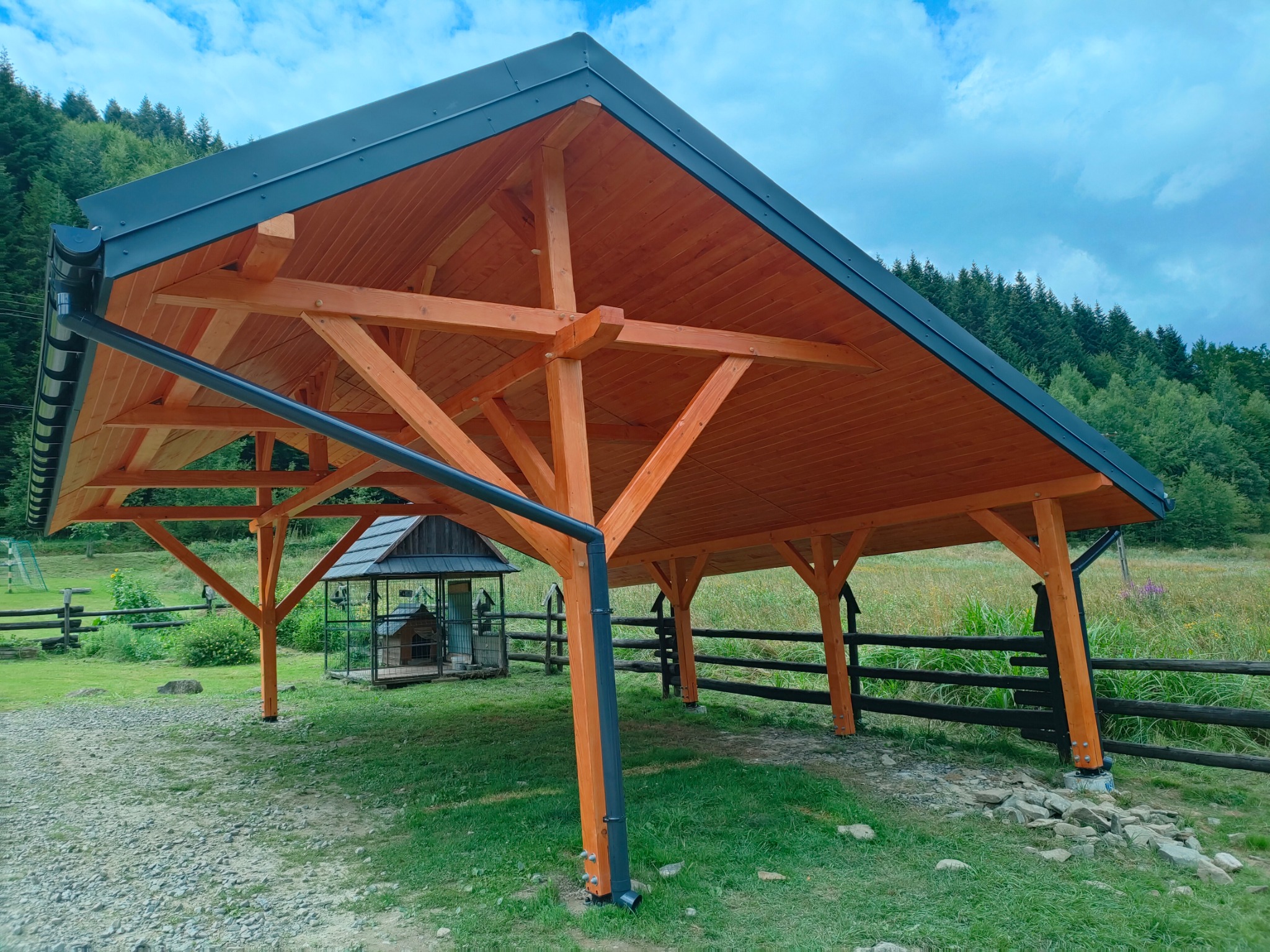 Otwarta, drewniana konstrukcja z ciemnym dachem i rynnami, na tle zieleni i błękitnego nieba. Widoczna drewniana podbudowa dachu i słupy nośne. W tle zagroda dla psa.