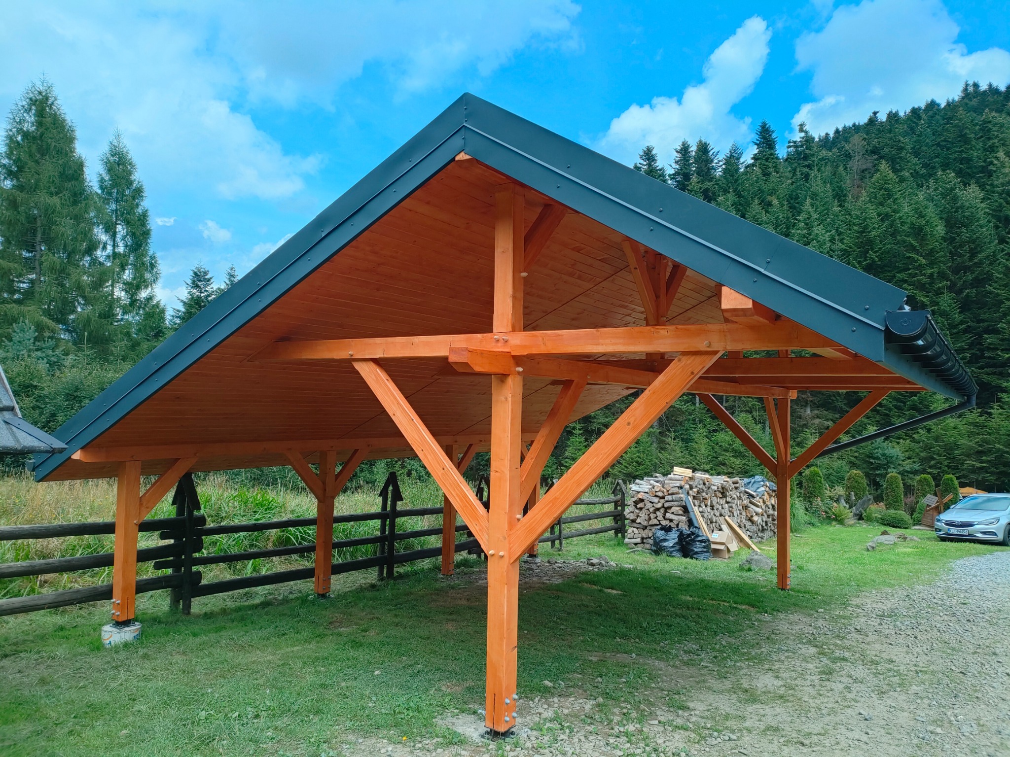 Nowoczesna, otwarta wiata z drewna w odcieniach pomarańczy, z ciemnym dachem na tle lasu i błękitnego nieba. Widoczne drewniane słupy i konstrukcja dachu.