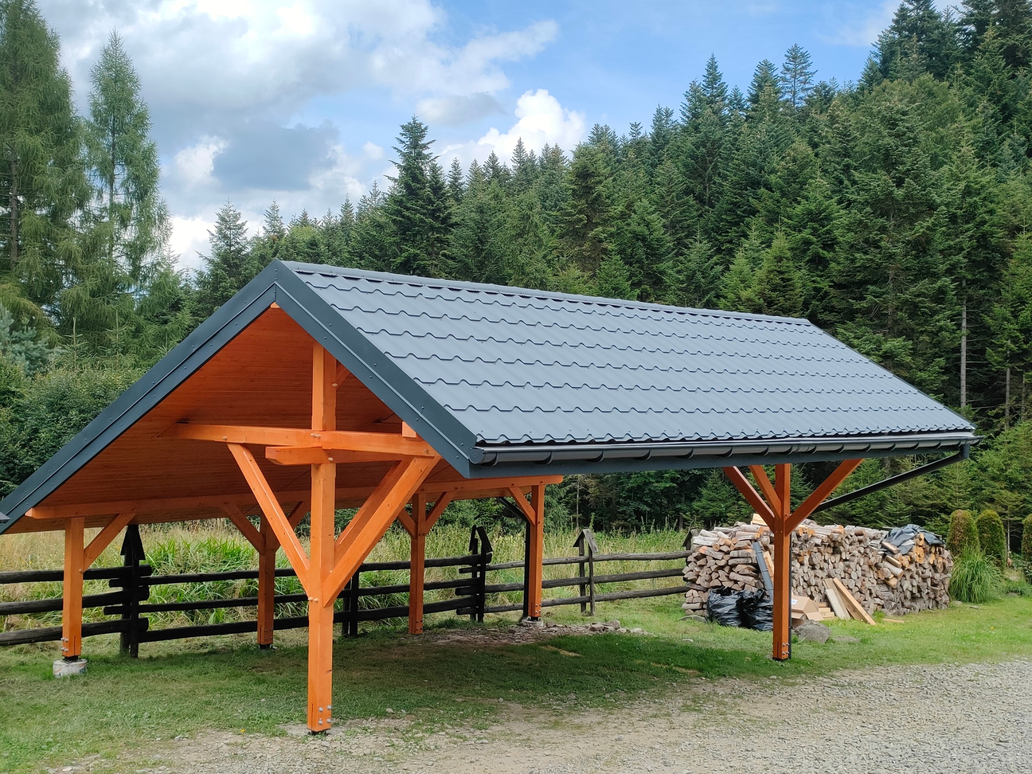 Drewniana wiata z szarą blachodachówką na tle lasu. Konstrukcja z belek w odcieniu miodowym. Obok ułożone drewno na opał. Widok na zewnątrz z wnętrza wiaty.