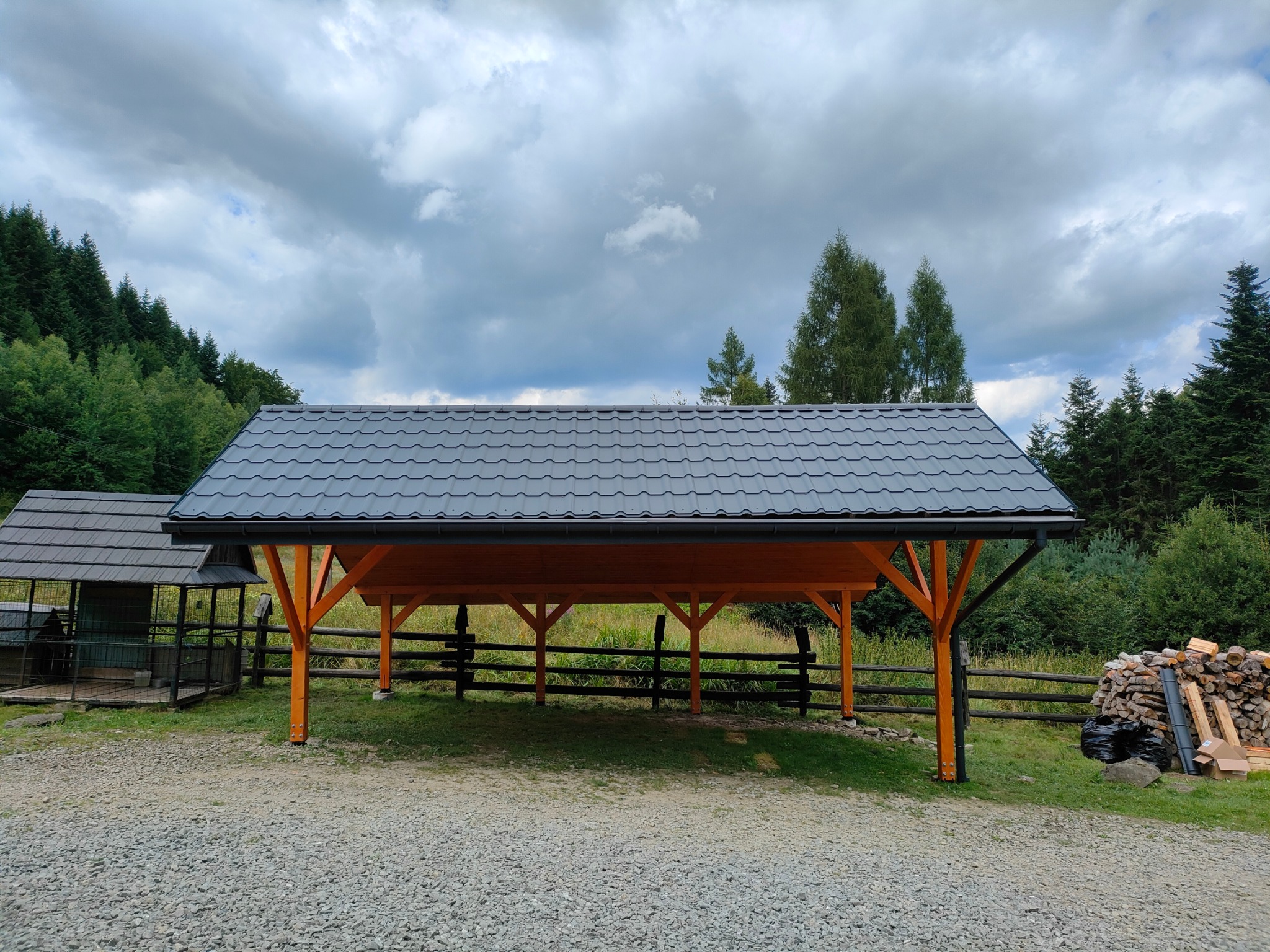 Zadaszenie z drewnianą konstrukcją i szarym dachem na tle zieleni. Widoczny fragment ogrodzenia i sterta drewna. Ujęcie z dołu, pochmurne niebo.
