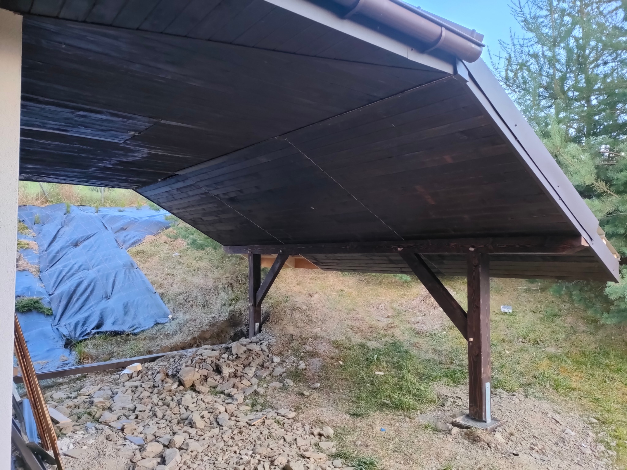 Ciemna, drewniana konstrukcja z dachem, wsparta na słupach, w trakcie budowy. W tle ziemia przykryta plandeką i drzewa. Widok z dołu, z boku.