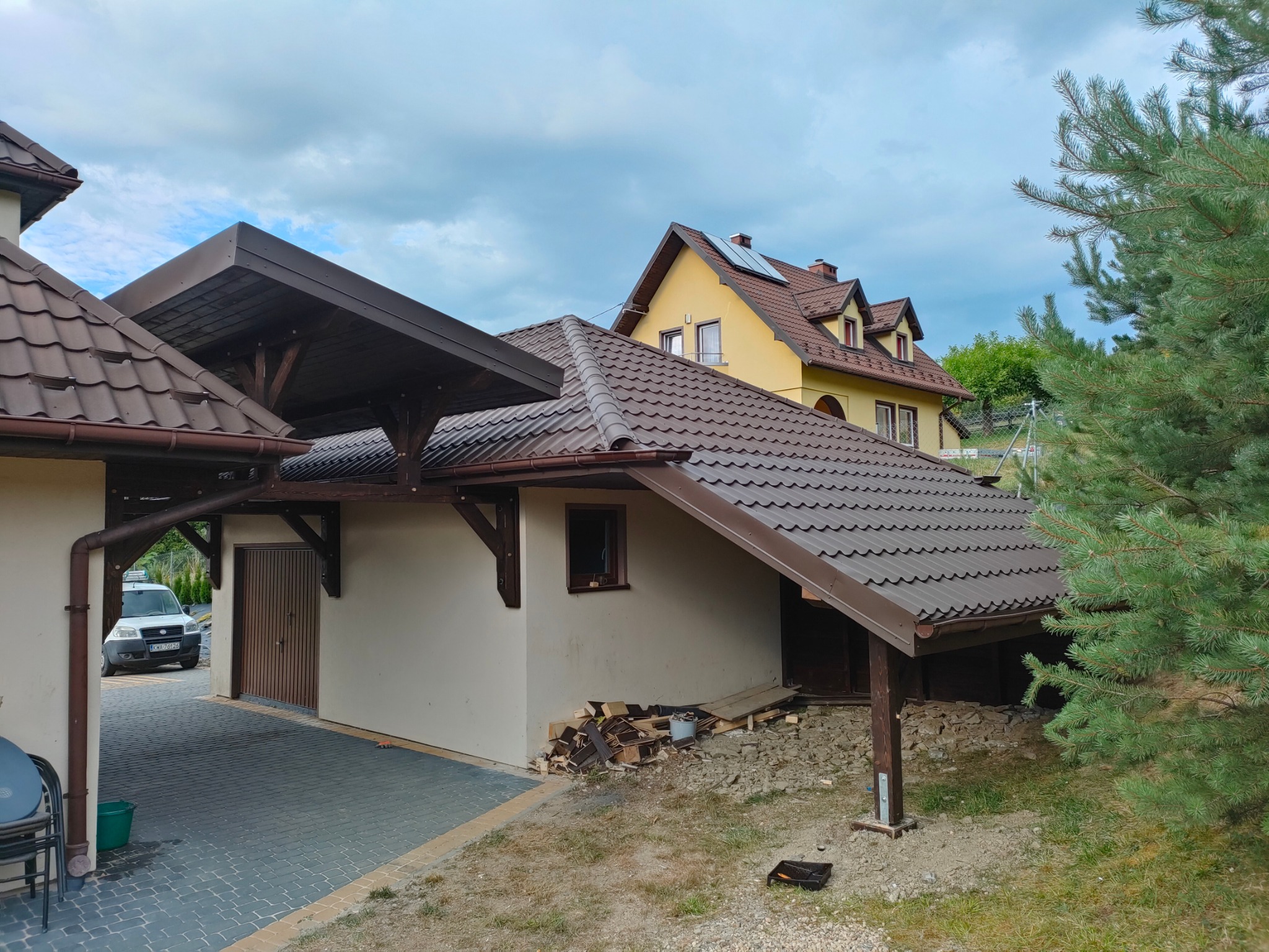 Zadaszenie z brązową dachówką nad wjazdem do garażu, widok z zewnątrz. W tle dom z żółtą elewacją i brązowym dachem. Częściowo widoczna zieleń drzew.