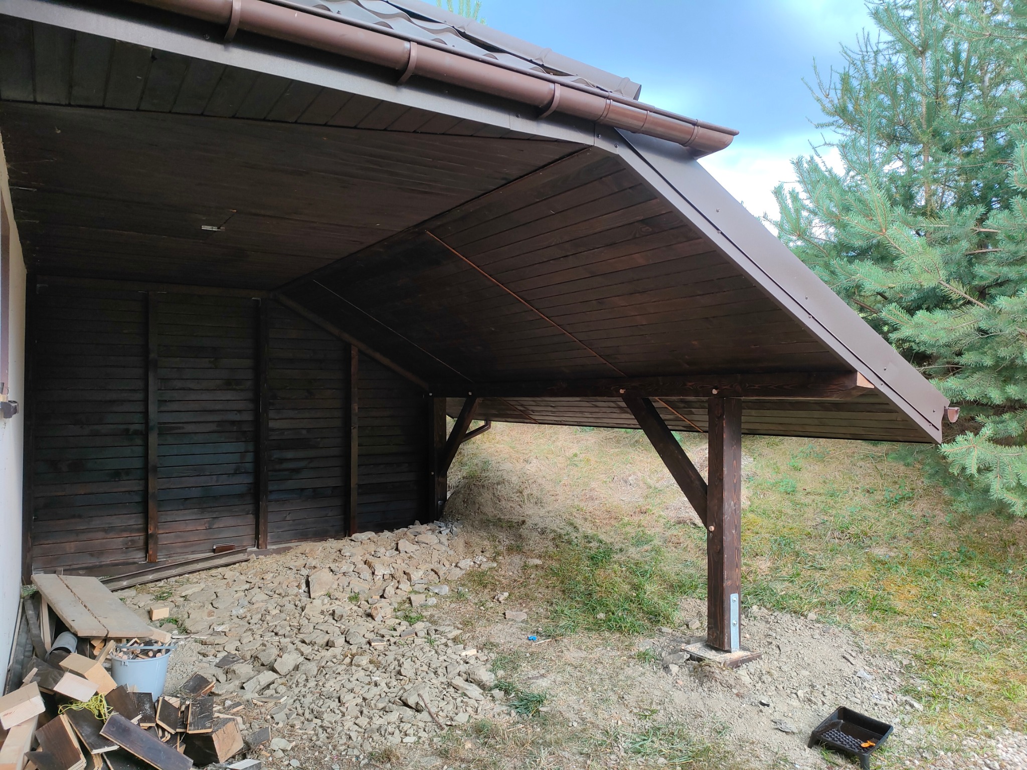 Ciemnobrązowa konstrukcja z drewna, prawdopodobnie zadaszenie, wsparta na słupach, z widocznymi elementami montażowymi. W tle zieleń i fragment budynku.