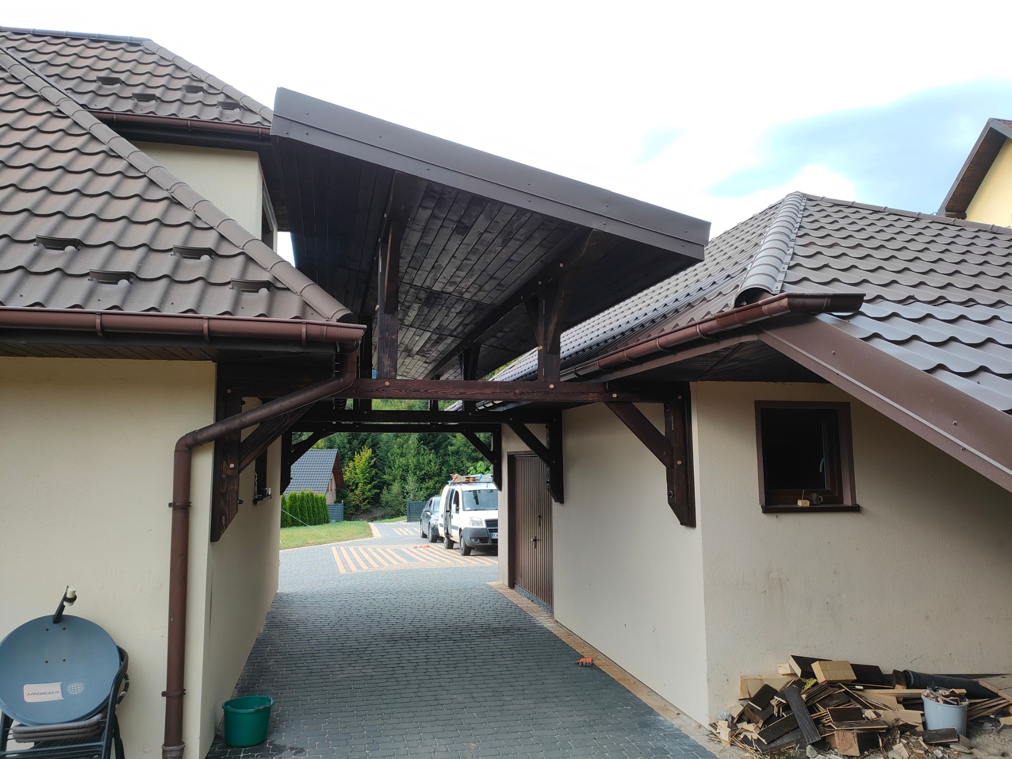 Drewniana pergola łącząca budynki z brązowym dachem, widok na podjazd z kostki brukowej i zaparkowane samochody. Widoczne drewniane elementy konstrukcyjne.