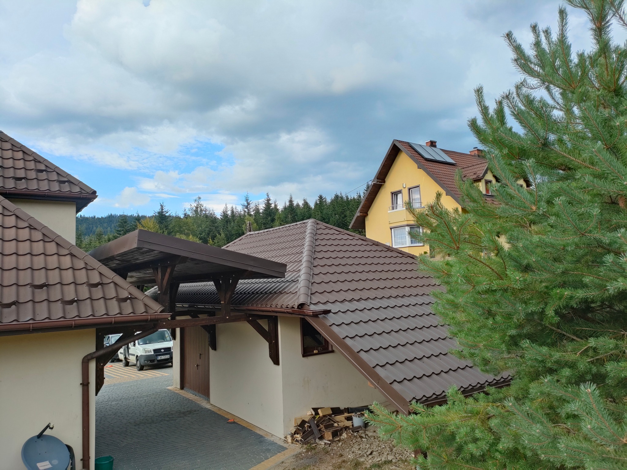 Zadaszenie nad wjazdem do garażu z brązową dachówką, wsparte na drewnianych belkach. W tle dom z panelami słonecznymi i zielone drzewa.