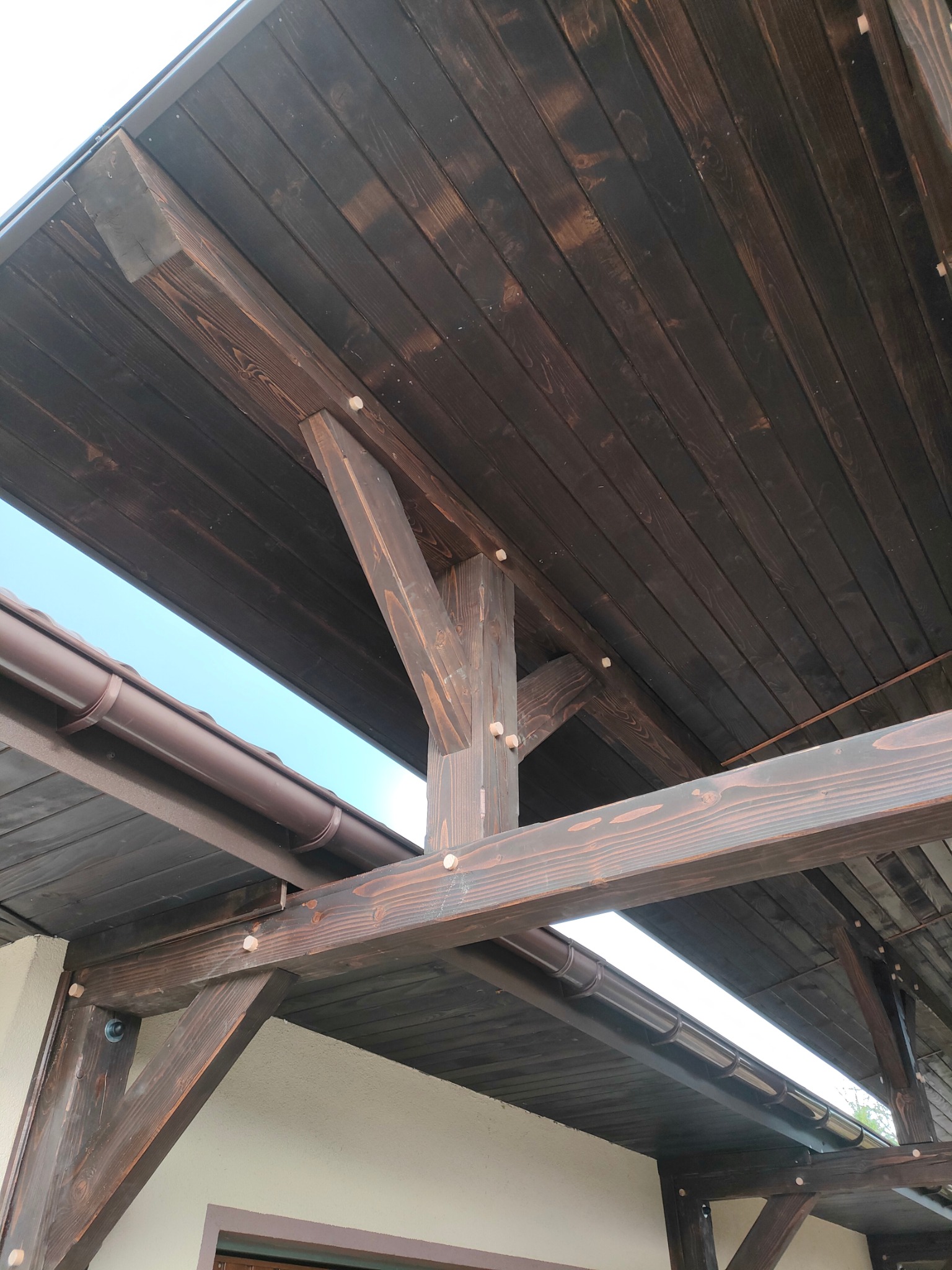 Ciemnobrązowa konstrukcja z drewna, prawdopodobnie zadaszenie tarasu lub werandy, z widocznymi elementami mocującymi i rynną. Ujęcie od dołu, widać fragment nieba.