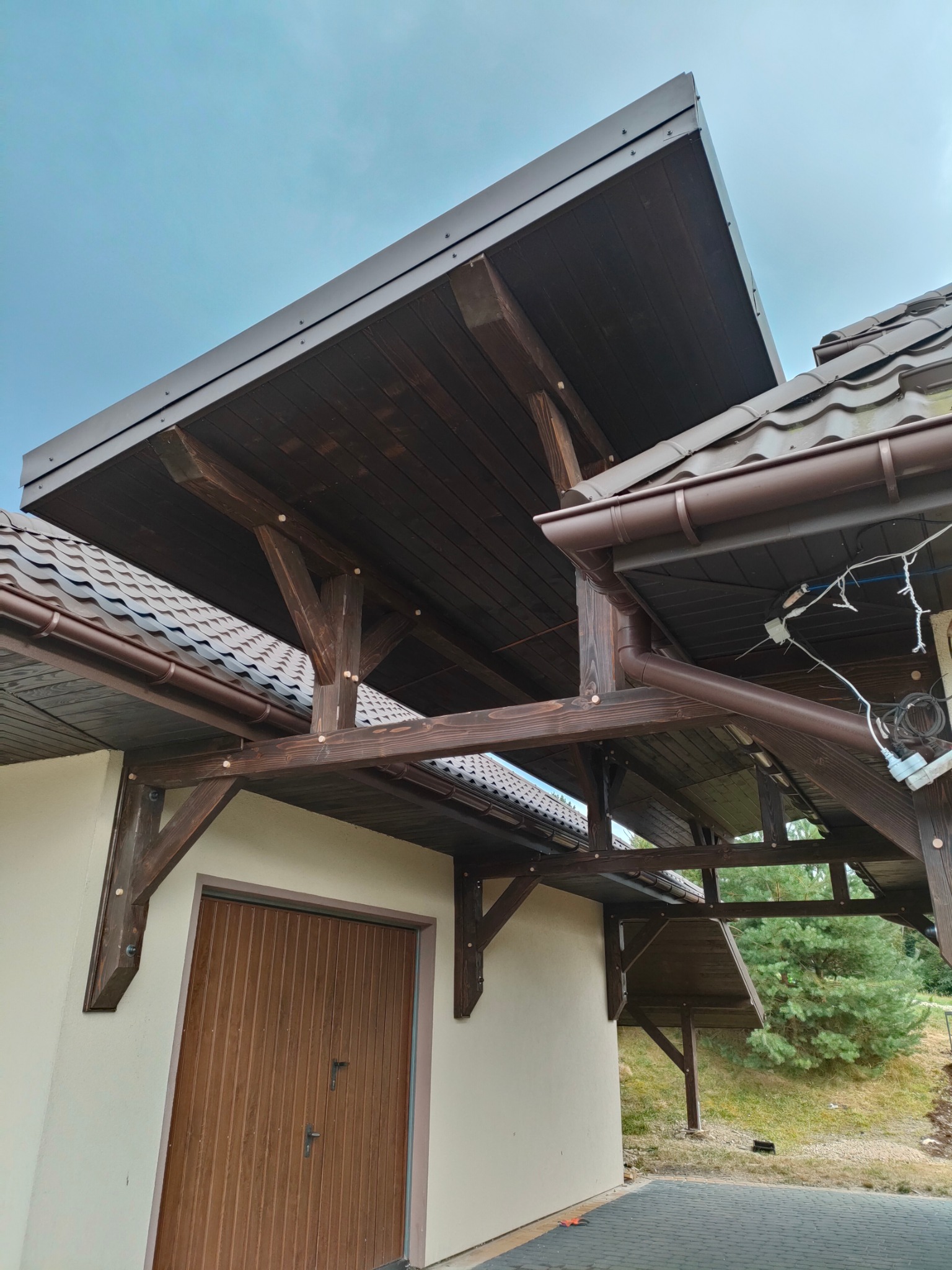 Ciemnobrązowa konstrukcja z drewna i metalu, prawdopodobnie zadaszenie nad wejściem lub tarasem, z widocznymi elementami dachu i rynny. Ujęcie z dołu, na tle pochmurnego nieba.