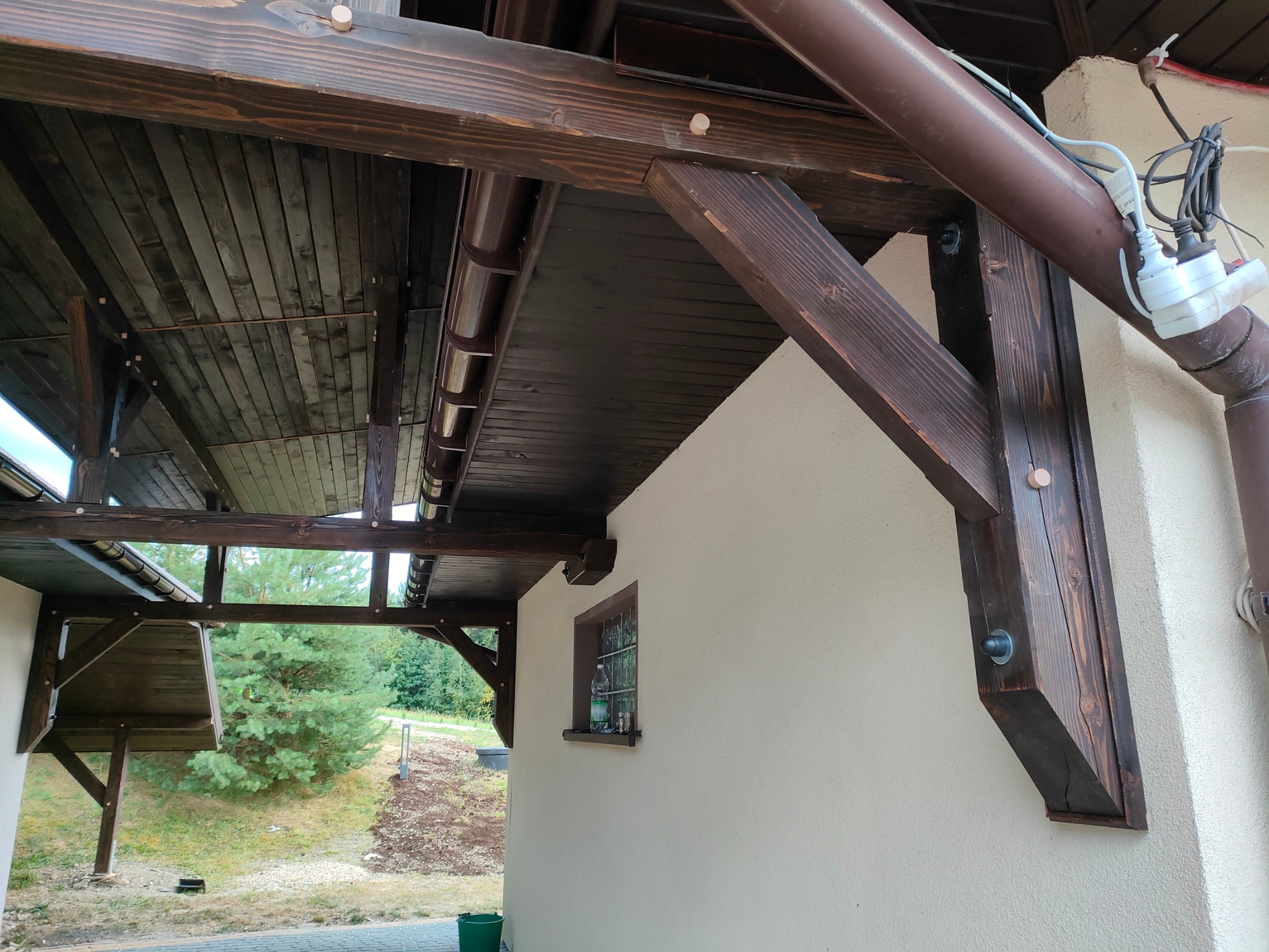 Zadaszenie z ciemnego drewna, wsparte na solidnych belkach, przylegające do budynku z małym oknem. Widok na zieleń w tle, z elementami instalacji elektrycznej.
