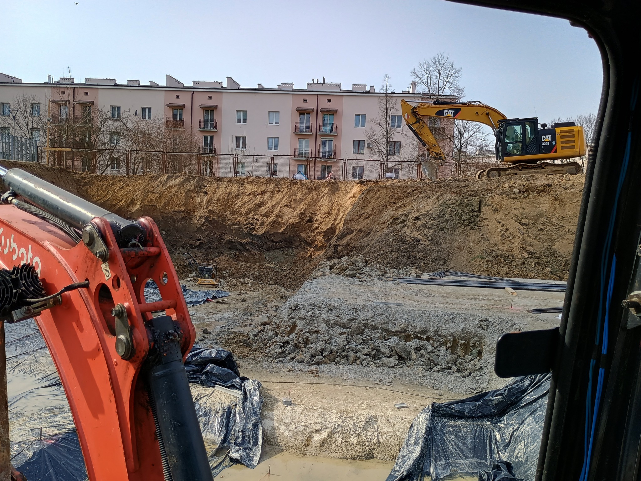 Widok z kabiny koparki Kubota na wykop z widoczną żółtą koparką CAT 320E na tle budynku mieszkalnego
