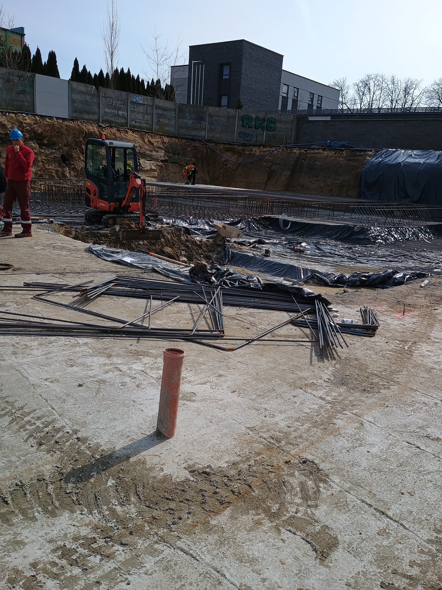 Przygotowanie terenu pod budowę: widoczny wykop zbrojony stalą, mała koparka, pracownicy w odblaskowych kamizelkach i kaskach, teren częściowo przykryty czarną folią, w tle budynki mieszkalne...