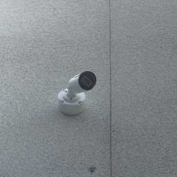 Sebastian Bolewski - Biała kamera monitoringu, zamontowana na szarej, teksturowanej ścianie. Widoczny kabel pionowy. Ujęcie z dołu, lekko z prawej strony.