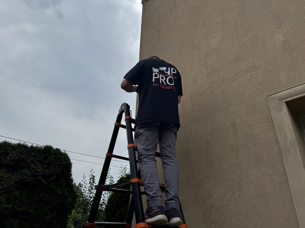 Instalator na drabinie montuje system alarmowy na elewacji domu. Widoczne logo firmy. Ujęcie z dołu, pochmurne niebo w tle.