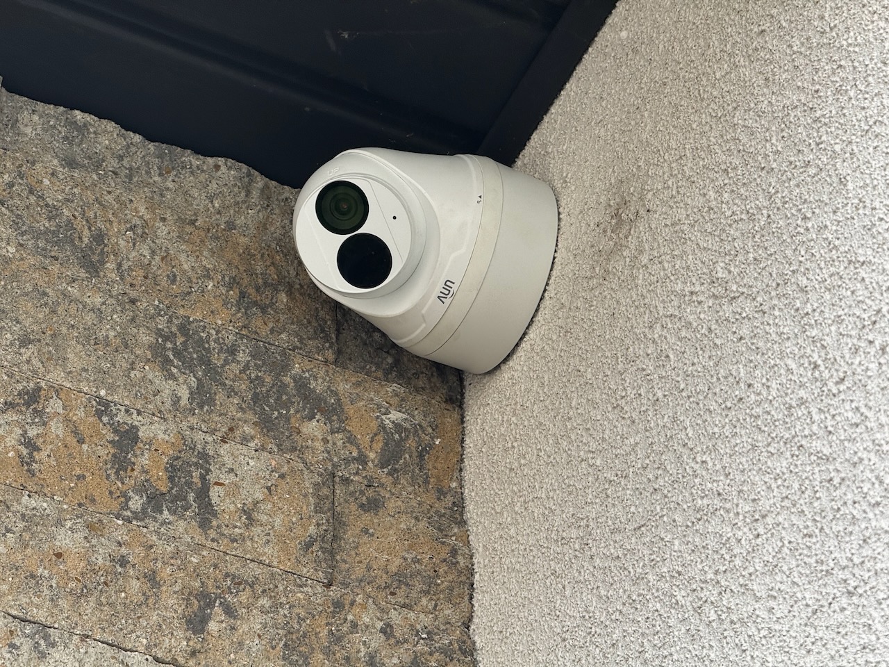 Biała kamera zewnętrzna AUN zamontowana na szorstkiej ścianie obok kamiennej elewacji, z widocznymi obiektywami i logo.