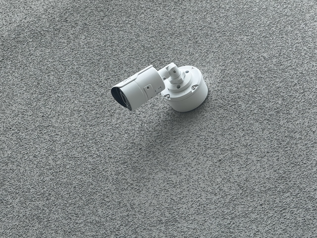Biała kamera monitoringu UNV zamontowana na szarej, chropowatej ścianie. Kąt ujęcia z dołu, skupienie na detalu mocowania i obudowy kamery.