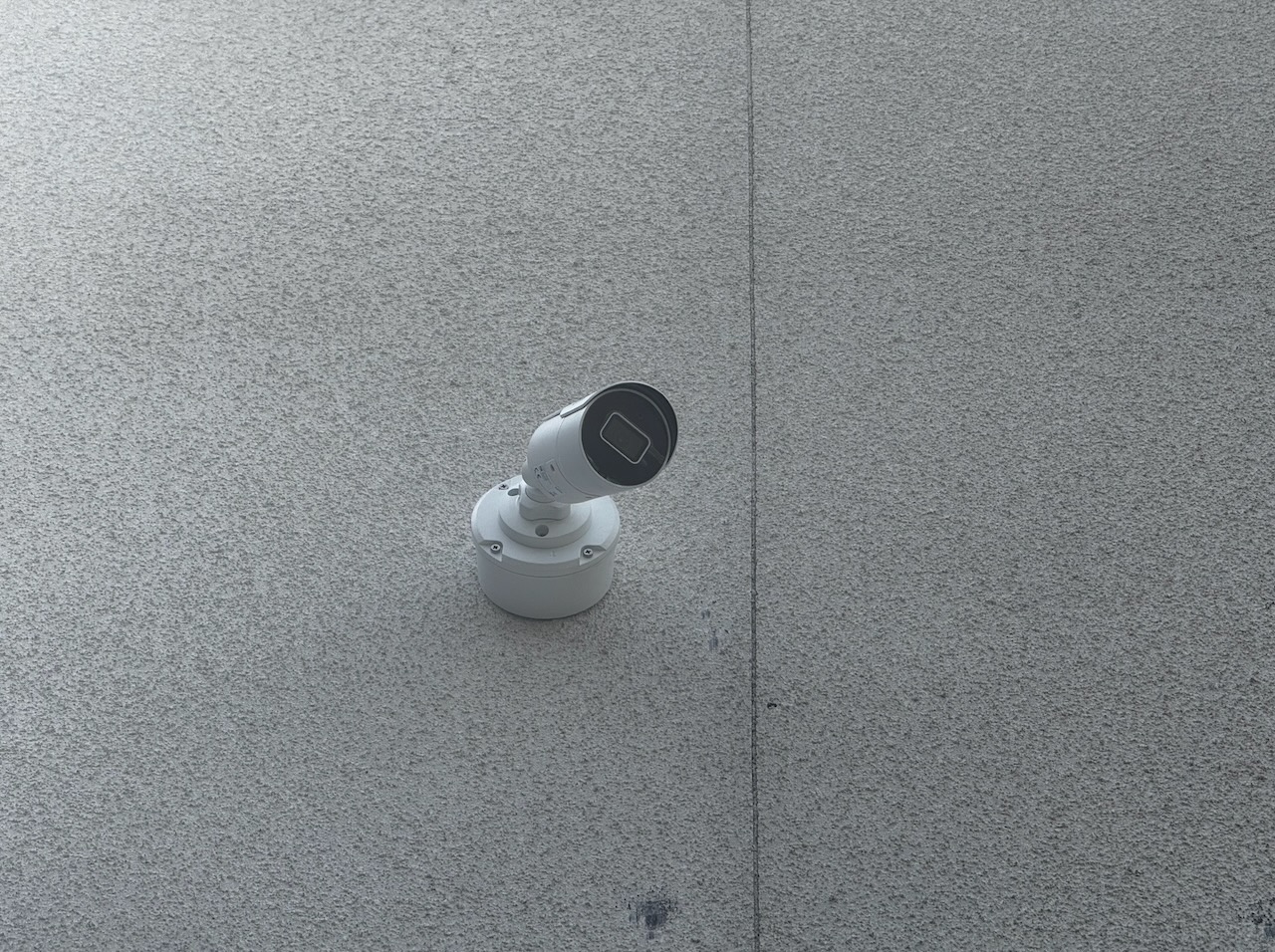 Biała kamera monitoringu, zamontowana na szarej, teksturowanej ścianie. Widoczny kabel pionowy. Ujęcie z dołu, lekko z prawej strony.