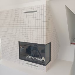 AS REMONTY SPÓŁKA Z OGRANICZONĄ ODPOWIEDZIALNOŚCIĄ - Nowoczesny kominek obudowany białymi, kwadratowymi płytkami, z widocznym paleniskiem i elementami dekoracyjnymi w postaci białych kamieni, w tle widoczny odkurzacz przemysłowy.