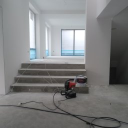 AS REMONTY SPÓŁKA Z OGRANICZONĄ ODPOWIEDZIALNOŚCIĄ - Wnętrze w trakcie malowania na biało, widoczne schody, okna zabezpieczone folią, agregat malarski Yato oraz puszka farby Magnat Perfect Matt.