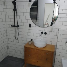 AS REMONTY SPÓŁKA Z OGRANICZONĄ ODPOWIEDZIALNOŚCIĄ - Wykończona łazienka z czarną armaturą, białą umywalką na drewnianej komodzie i białymi płytkami typu metro, okrągłe lustro z wiszącym oświetleniem, widoczny odpływ liniowy.