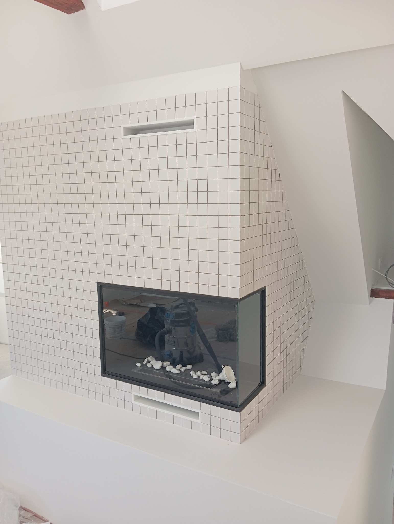 Nowoczesny kominek obudowany białymi, kwadratowymi płytkami, z widocznym paleniskiem i elementami dekoracyjnymi w postaci białych kamieni, w tle widoczny odkurzacz przemysłowy.