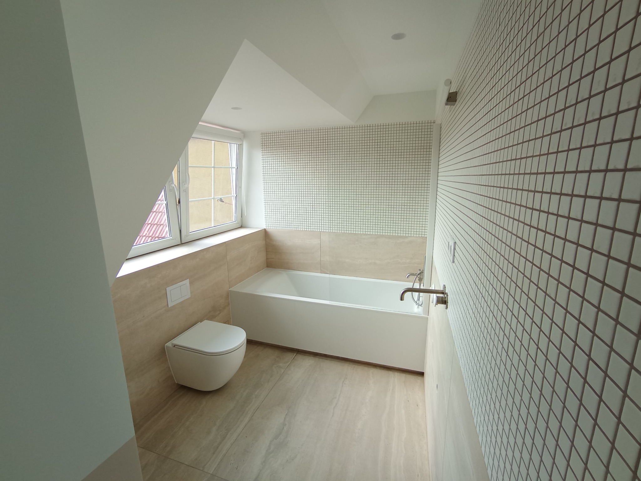 Nowoczesna łazienka na poddaszu z białymi płytkami mozaikowymi, podwieszaną toaletą i wanną, wykończona w beżowych odcieniach.