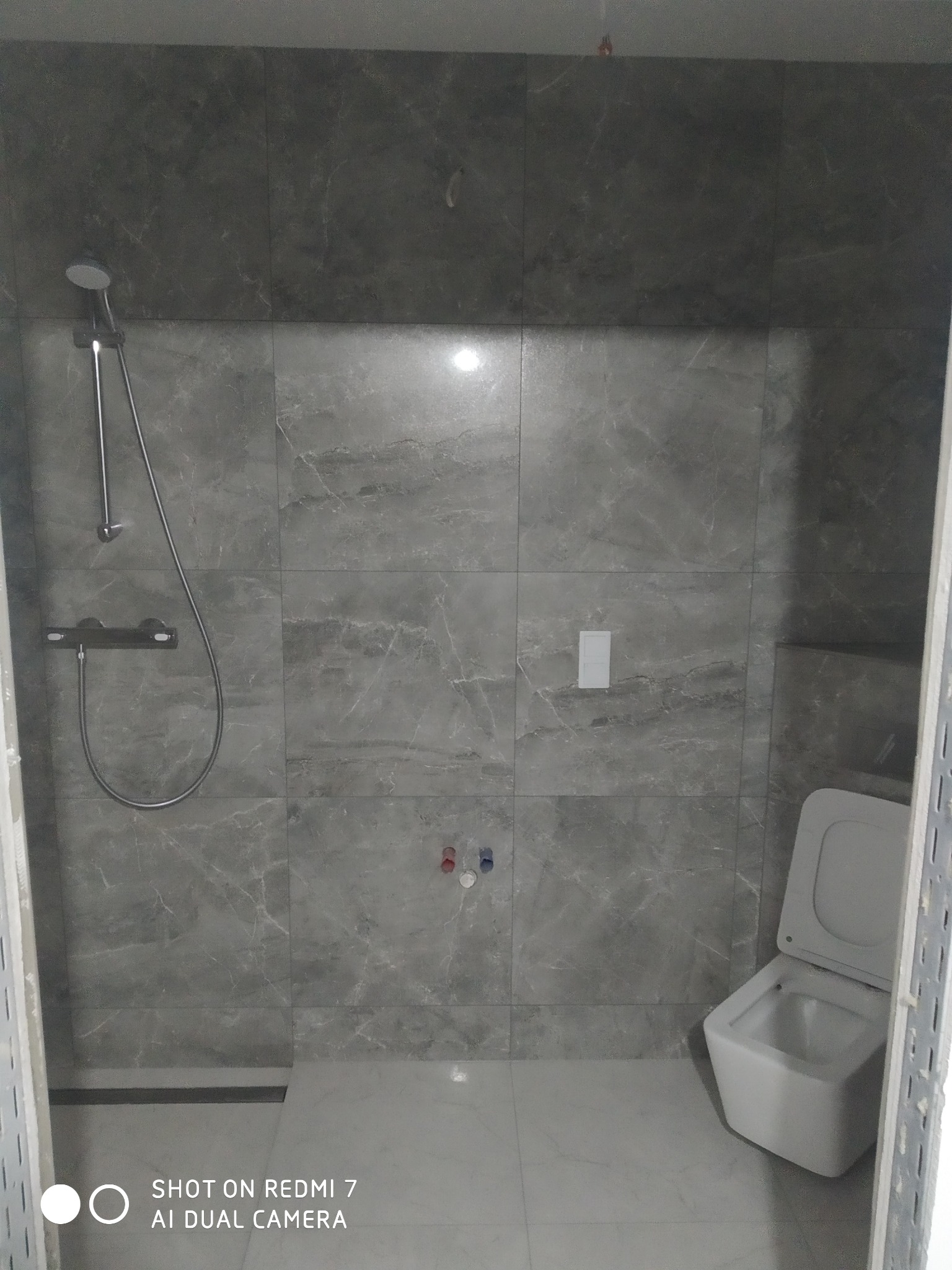 Nowoczesna kabina prysznicowa z szarymi płytkami imitującymi marmur, widoczna bateria, deszczownica i toaleta podwieszana z otwartą klapą, surowy stan wykończenia.