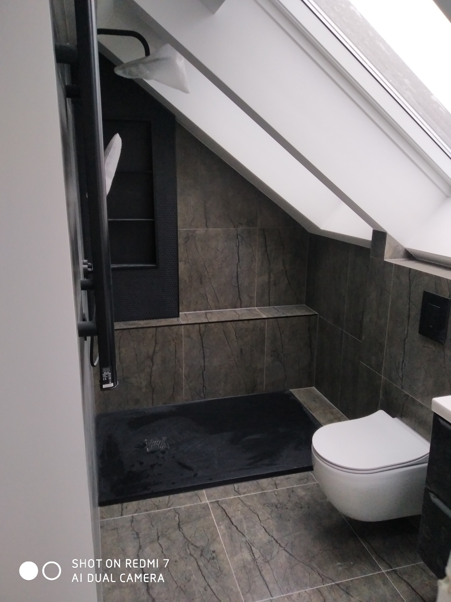 Nowoczesna łazienka na poddaszu w Gdańsku z kabiną prysznicową typu walk-in z czarnym brodzikiem, toaletą podwieszaną i płytkami imitującymi marmur.