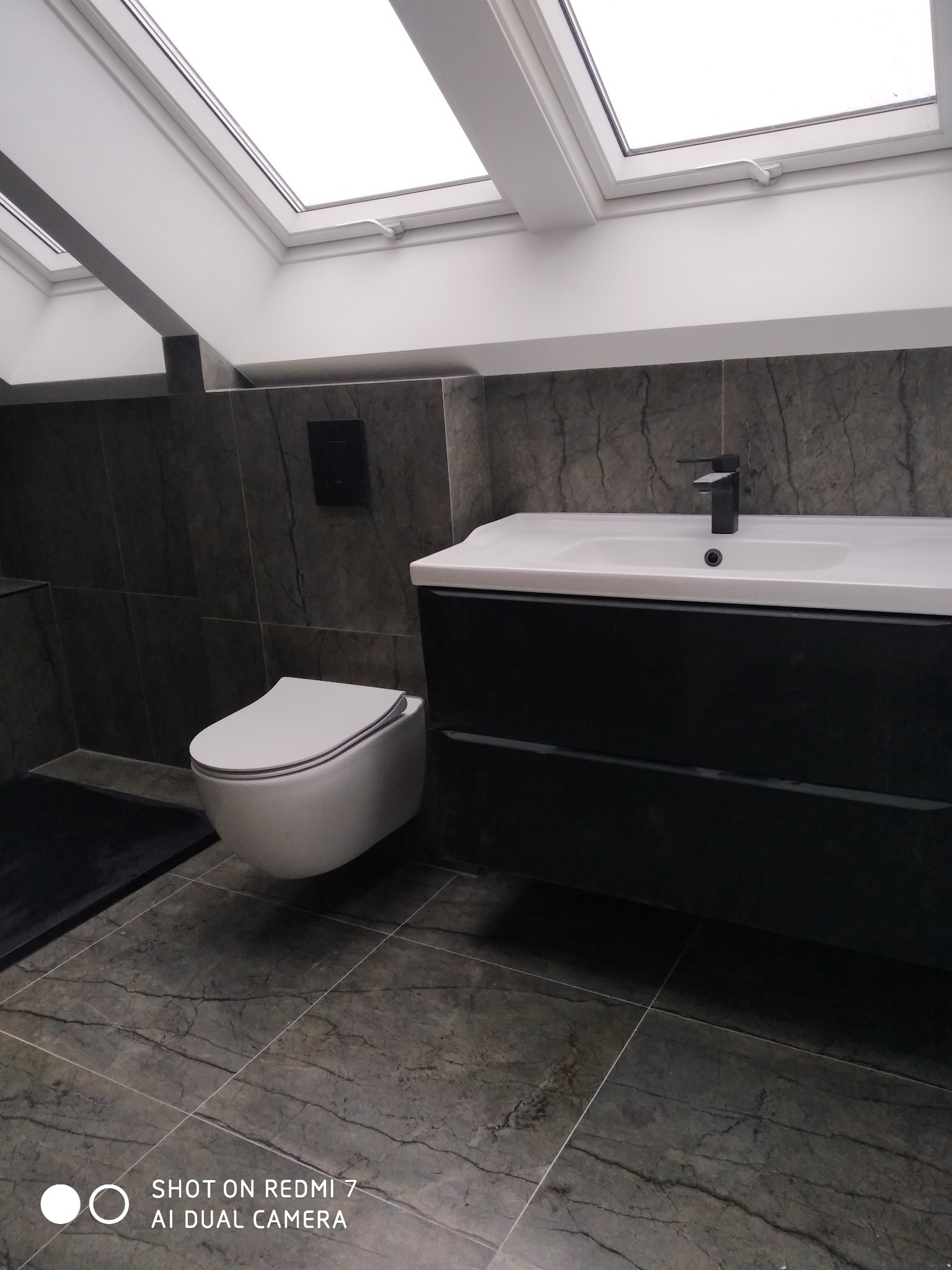 Nowoczesna łazienka z marmurowymi płytkami, podwieszaną toaletą i szafką z umywalką, doświetlona oknami dachowymi.