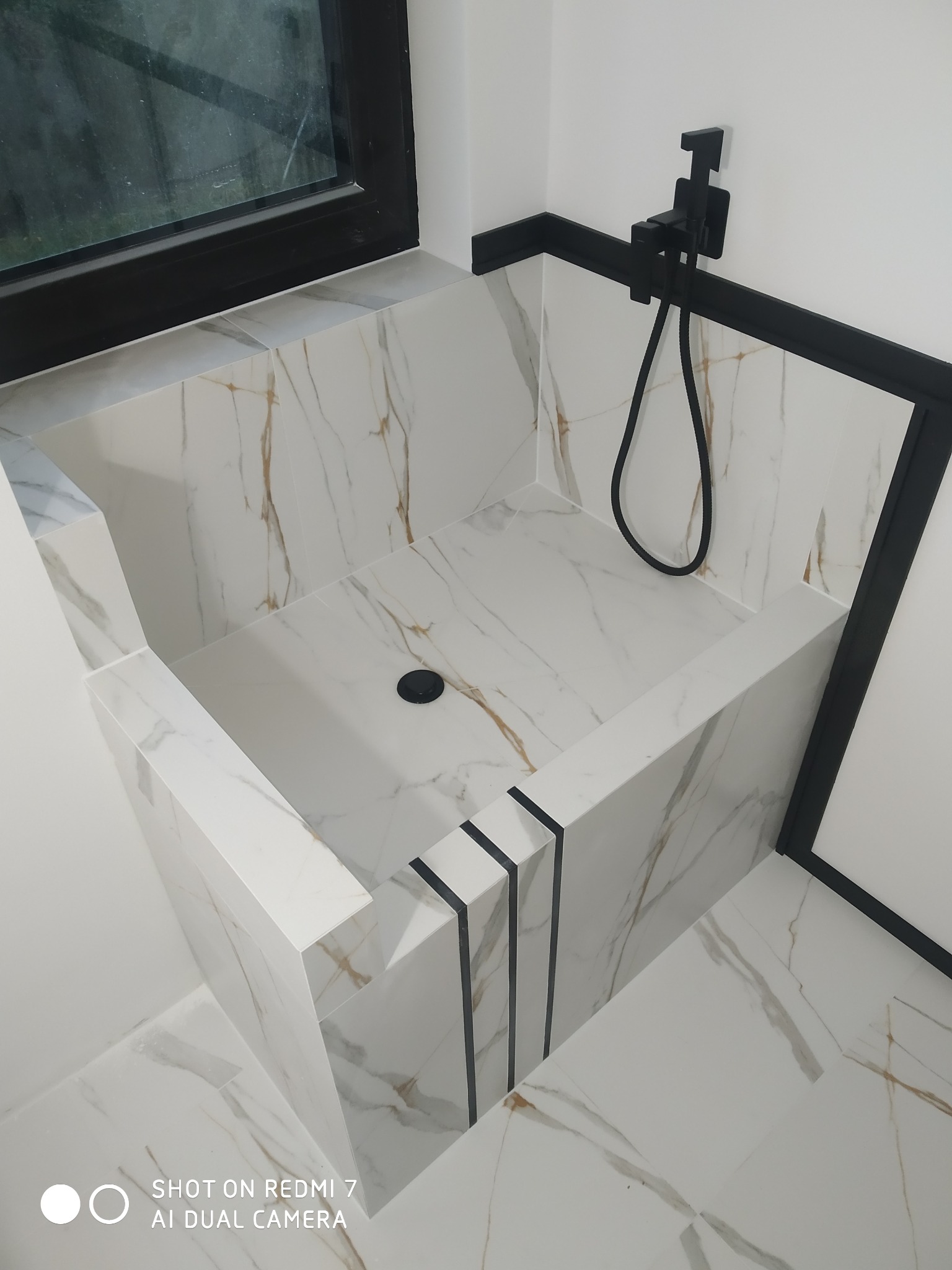 Nowoczesny brodzik wyłożony białymi płytkami z marmurkowym wzorem, czarna armatura i obramowanie kabiny, minimalistyczny design.