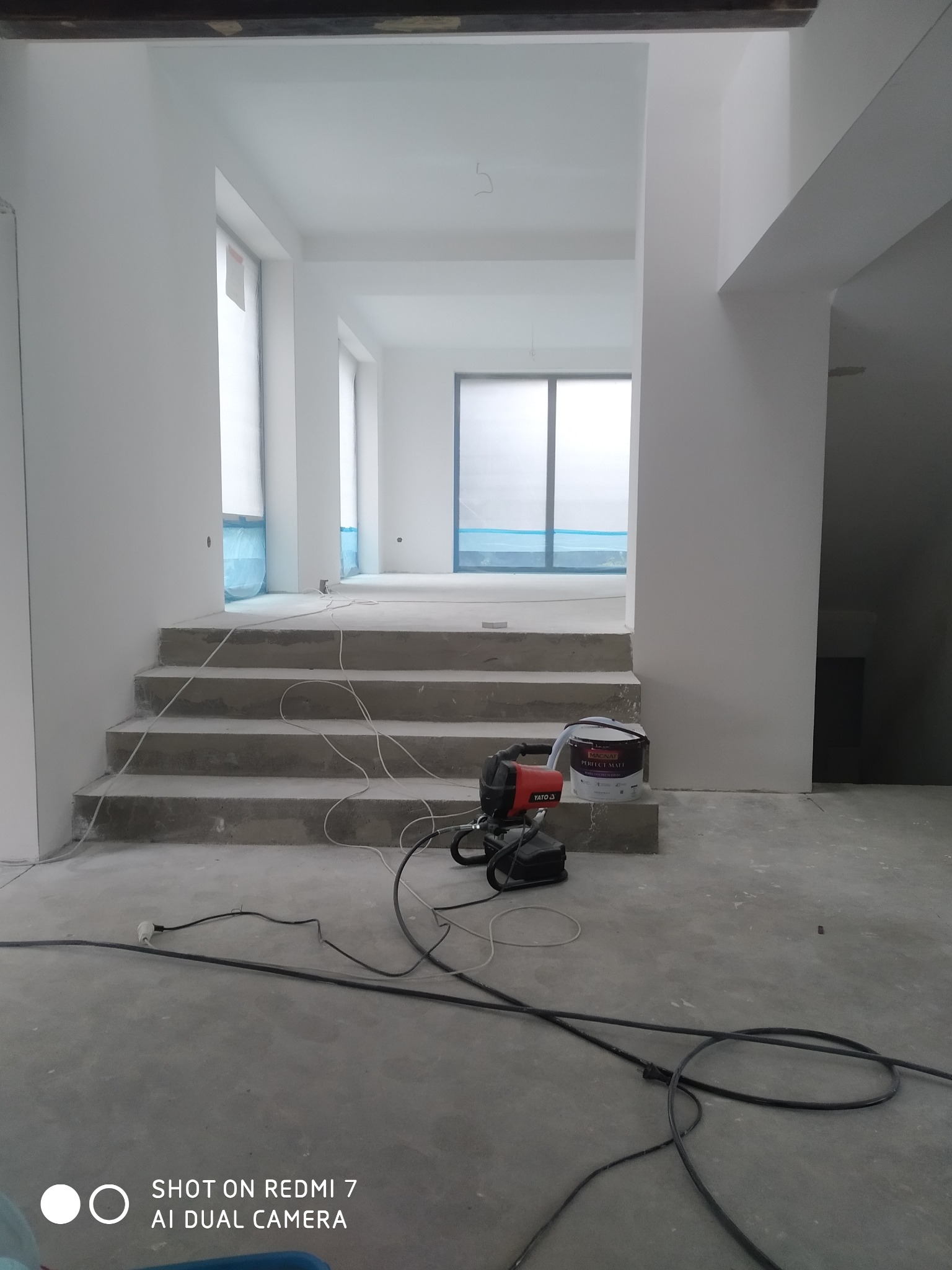 Wnętrze w trakcie malowania na biało, widoczne schody, okna zabezpieczone folią, agregat malarski Yato oraz puszka farby Magnat Perfect Matt.