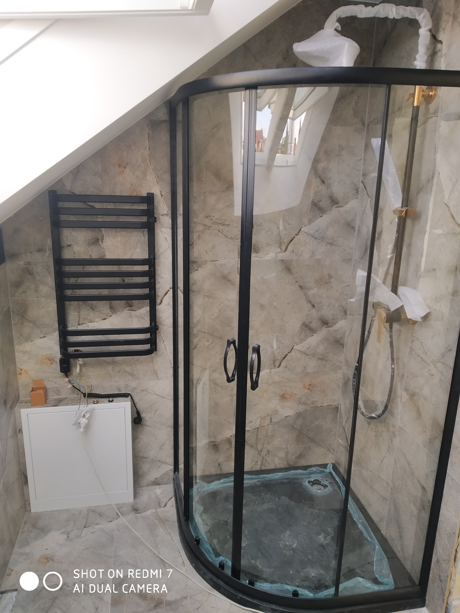 Czarna kabina prysznicowa z zaokrąglonymi szybami w łazience na poddaszu, z marmurowymi płytkami na ścianach i grzejnikiem drabinkowym, bateria i deszczownica zabezpieczone folią.