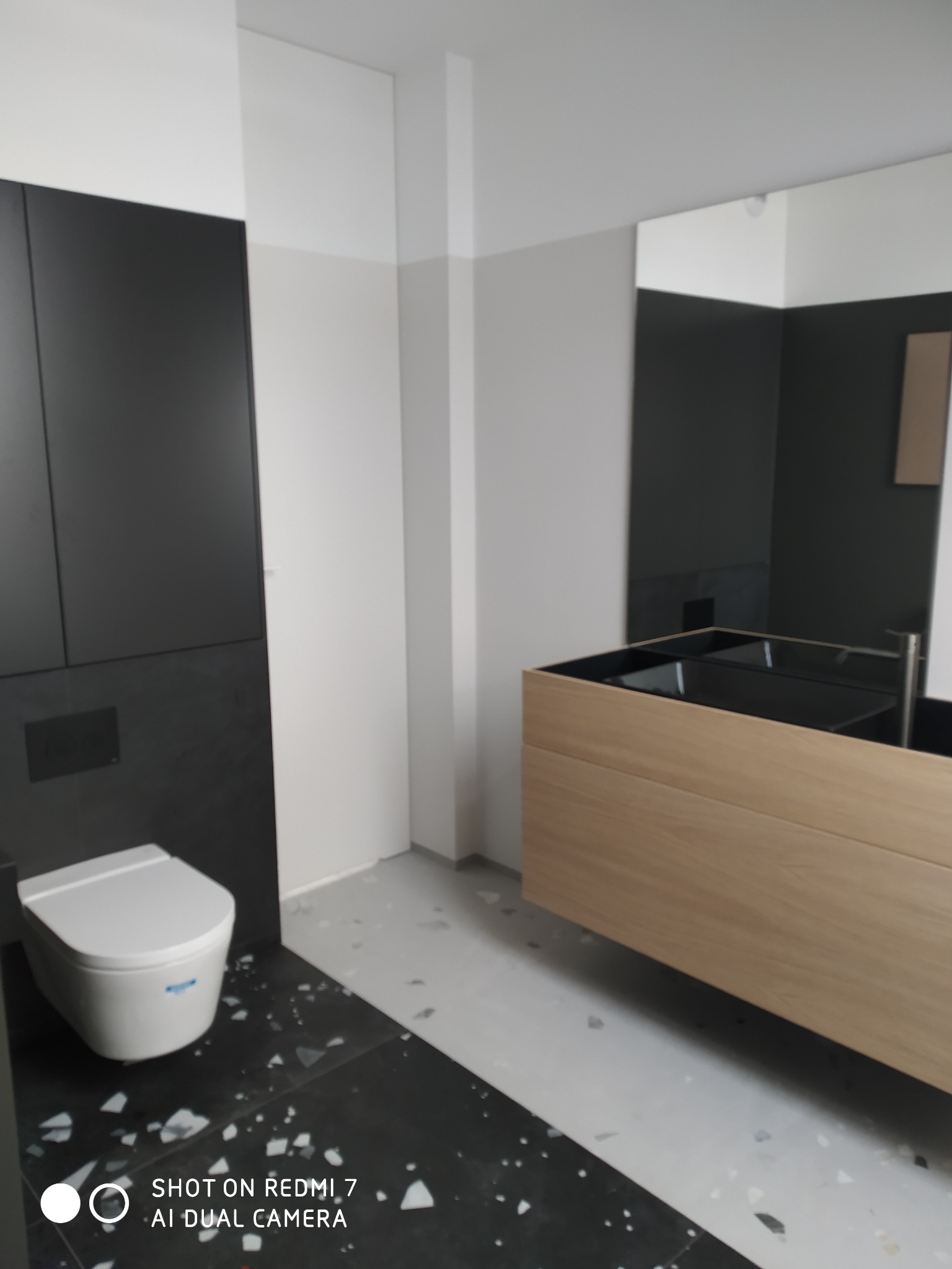 Nowoczesna łazienka w trakcie wykańczania: czarne płytki podłogowe z białymi elementami, podwieszana toaleta, drewniana szafka z czarną umywalką, lustro i częściowo pomalowane ściany.