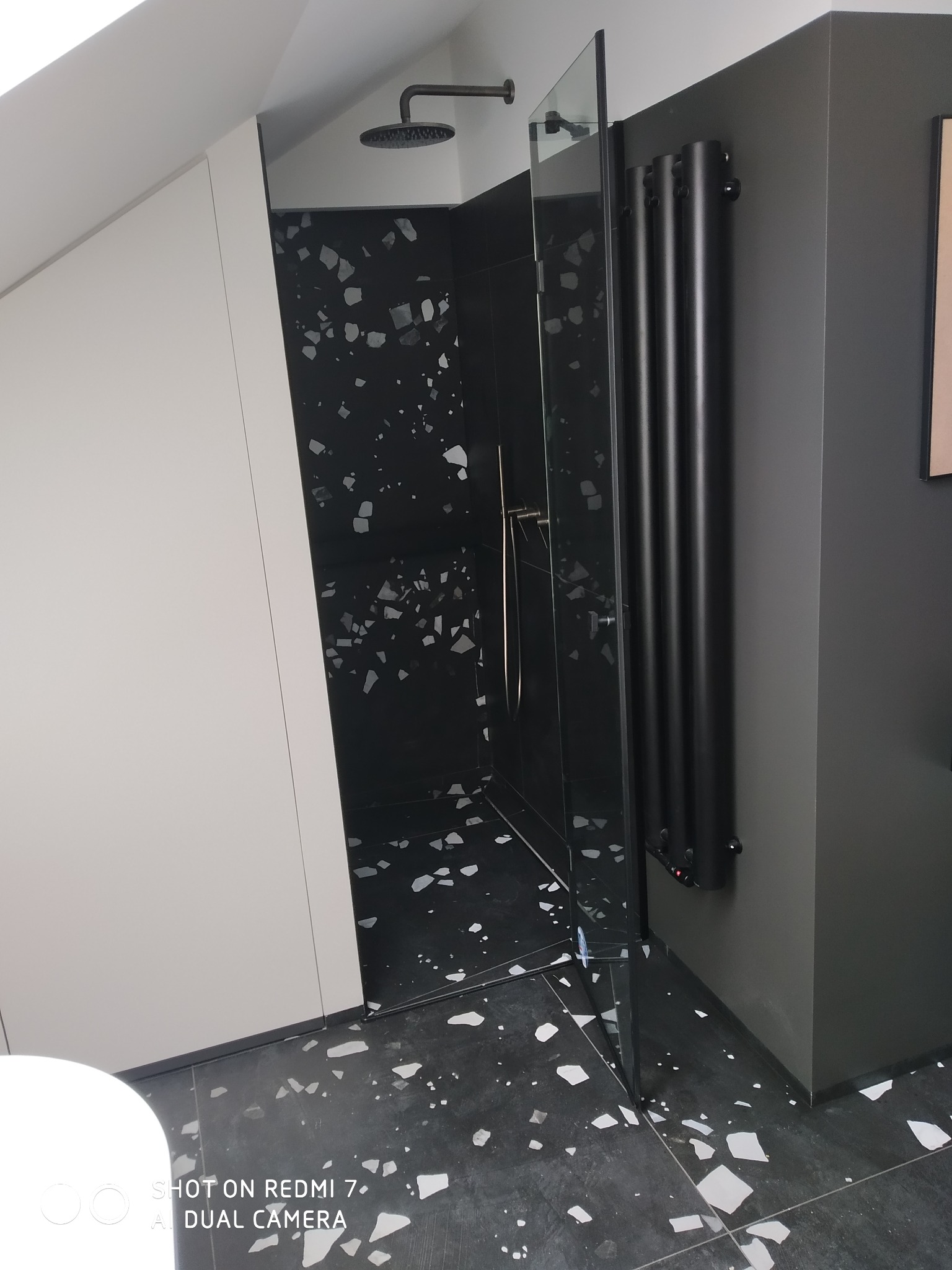 Nowoczesna łazienka w odcieniach czerni i bieli, z kabiną prysznicową typu walk-in, czarną armaturą i grzejnikiem drabinkowym, wykończona płytkami imitującymi lastryko.