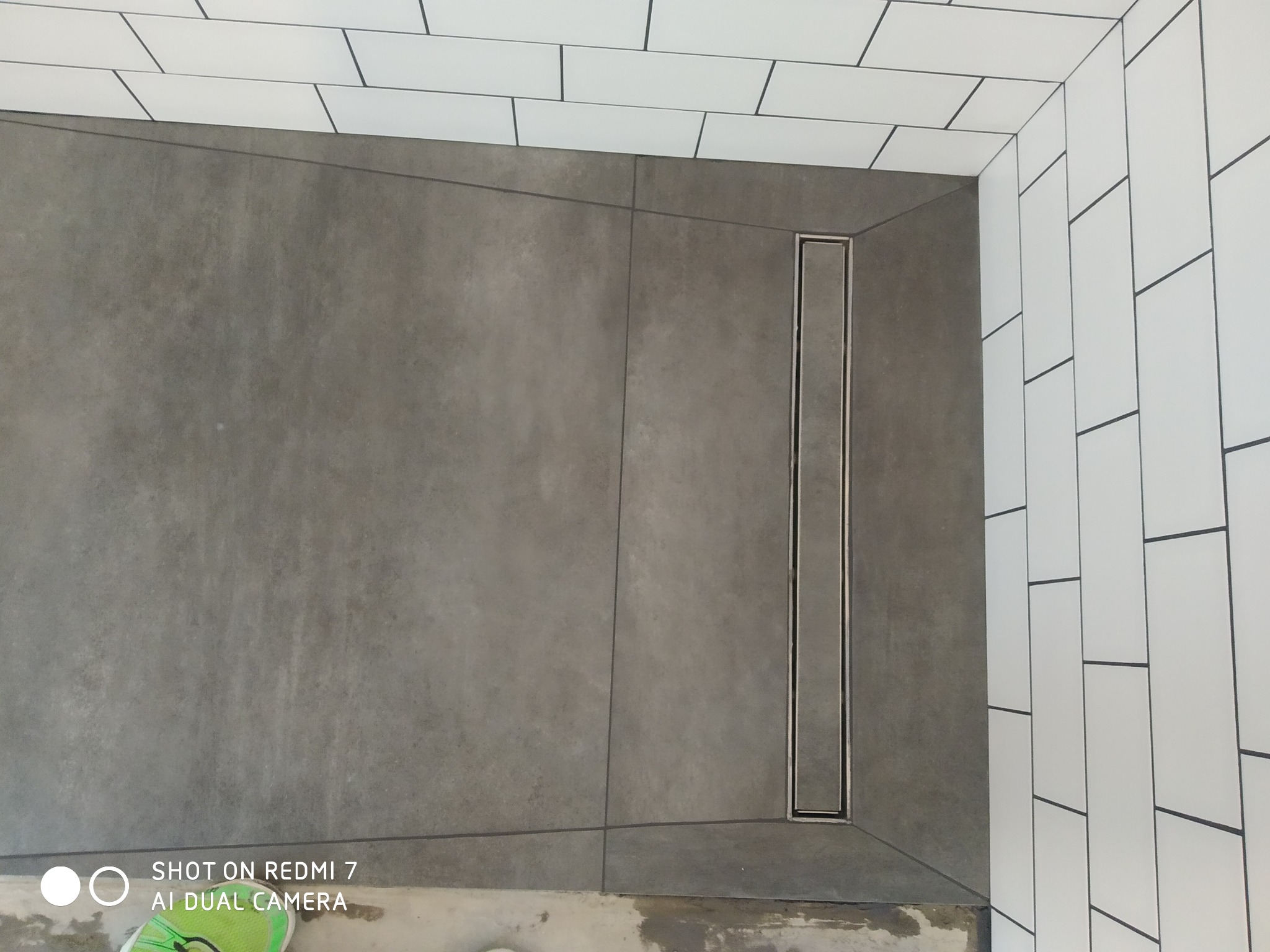 Szara posadzka prysznicowa z odpływem liniowym i białe, prostokątne płytki ścienne z ciemną fugą, widoczne fragmenty podłoża w trakcie prac.