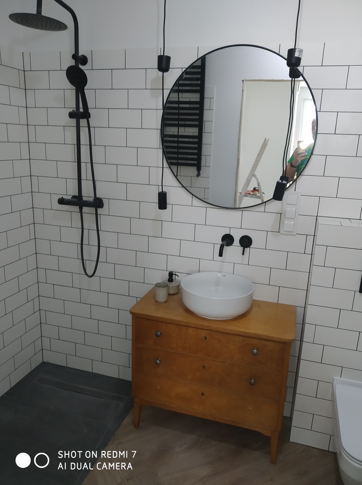 Wykończona łazienka z czarną armaturą, białą umywalką na drewnianej komodzie i białymi płytkami typu metro, okrągłe lustro z wiszącym oświetleniem, widoczny odpływ liniowy.