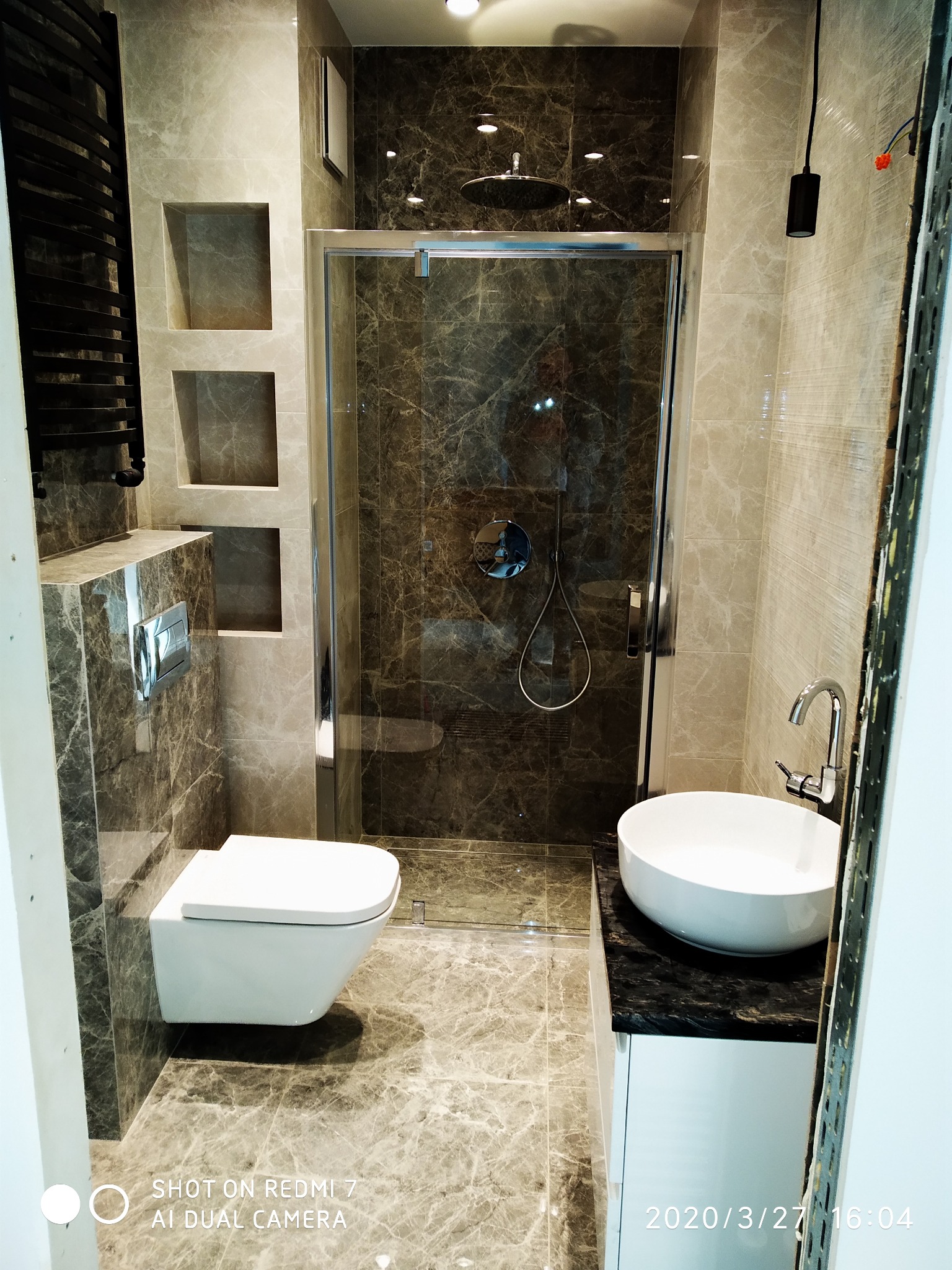 Wykończona łazienka z marmurowymi płytkami, kabina prysznicowa typu walk-in z deszczownicą, podwieszana toaleta i umywalka nablatowa na czarnym blacie, wnęki w ścianie.