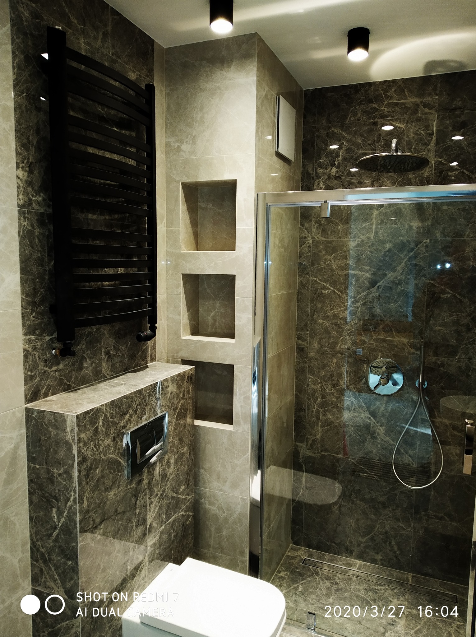 Wykończona łazienka w Gdańsku z marmurowymi płytkami, czarnym grzejnikiem drabinkowym, wnękami w ścianie i kabiną prysznicową typu walk-in z deszczownicą.